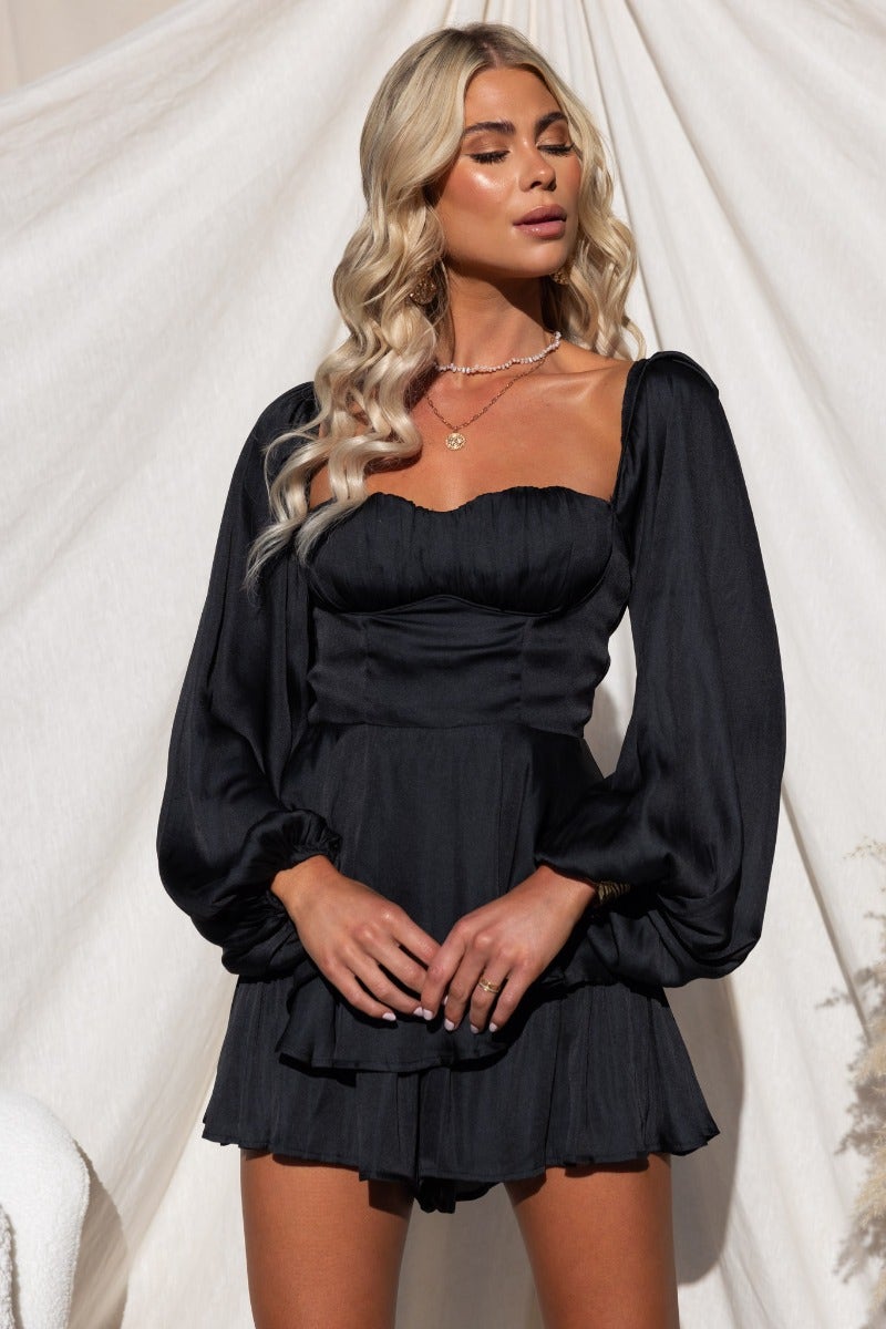 Model wearing a black Helly playsuit with padded bust and elastic sleeves, styled with a layered necklace.