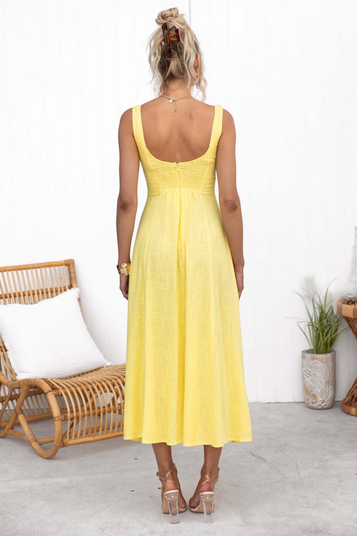 Back view of a woman in a yellow midi dress, showcasing faux buttons and a lined bust, styled with a casual setting.