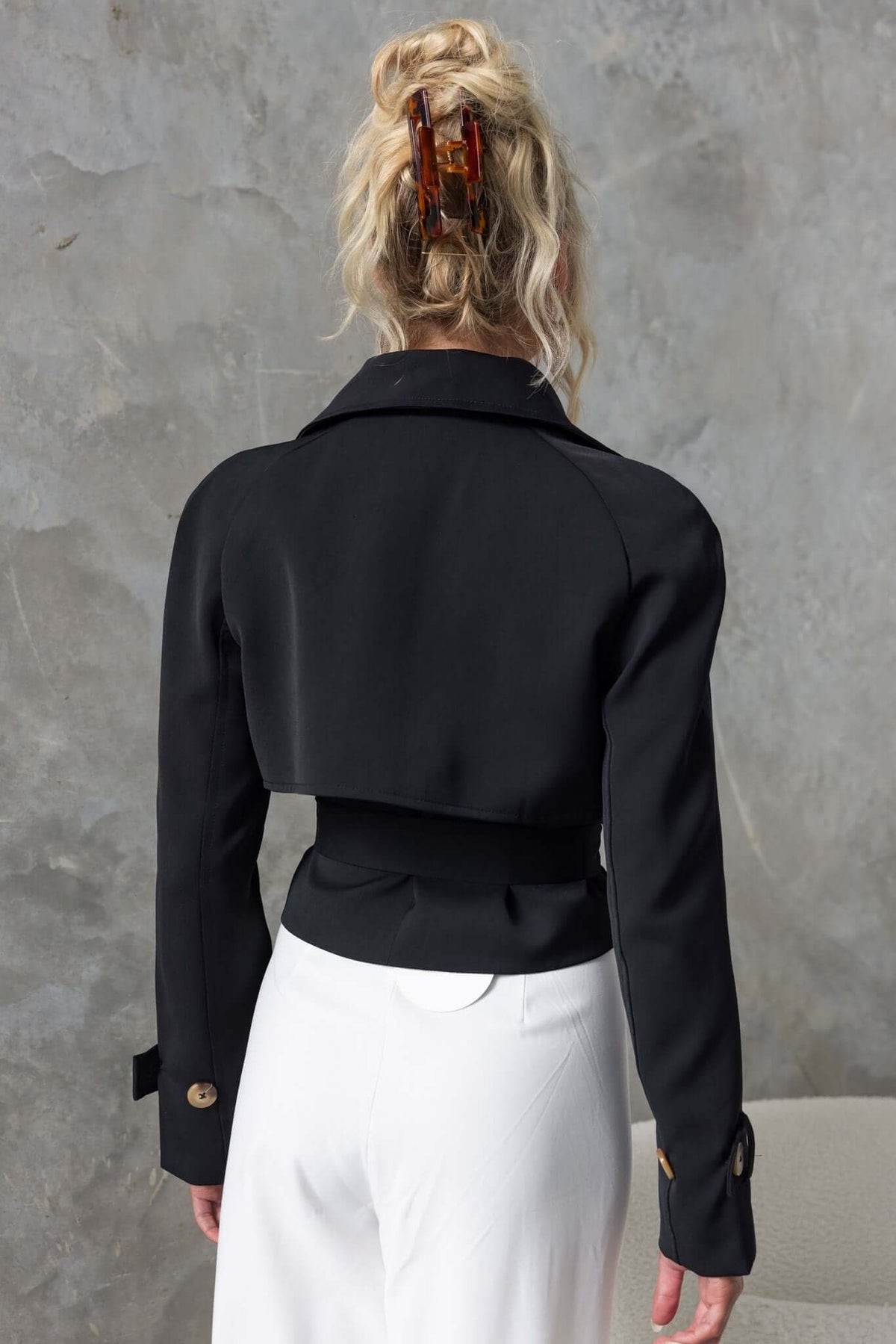 Back view of a cropped black trench coat featuring a collar and removable tie, styled with white trousers.