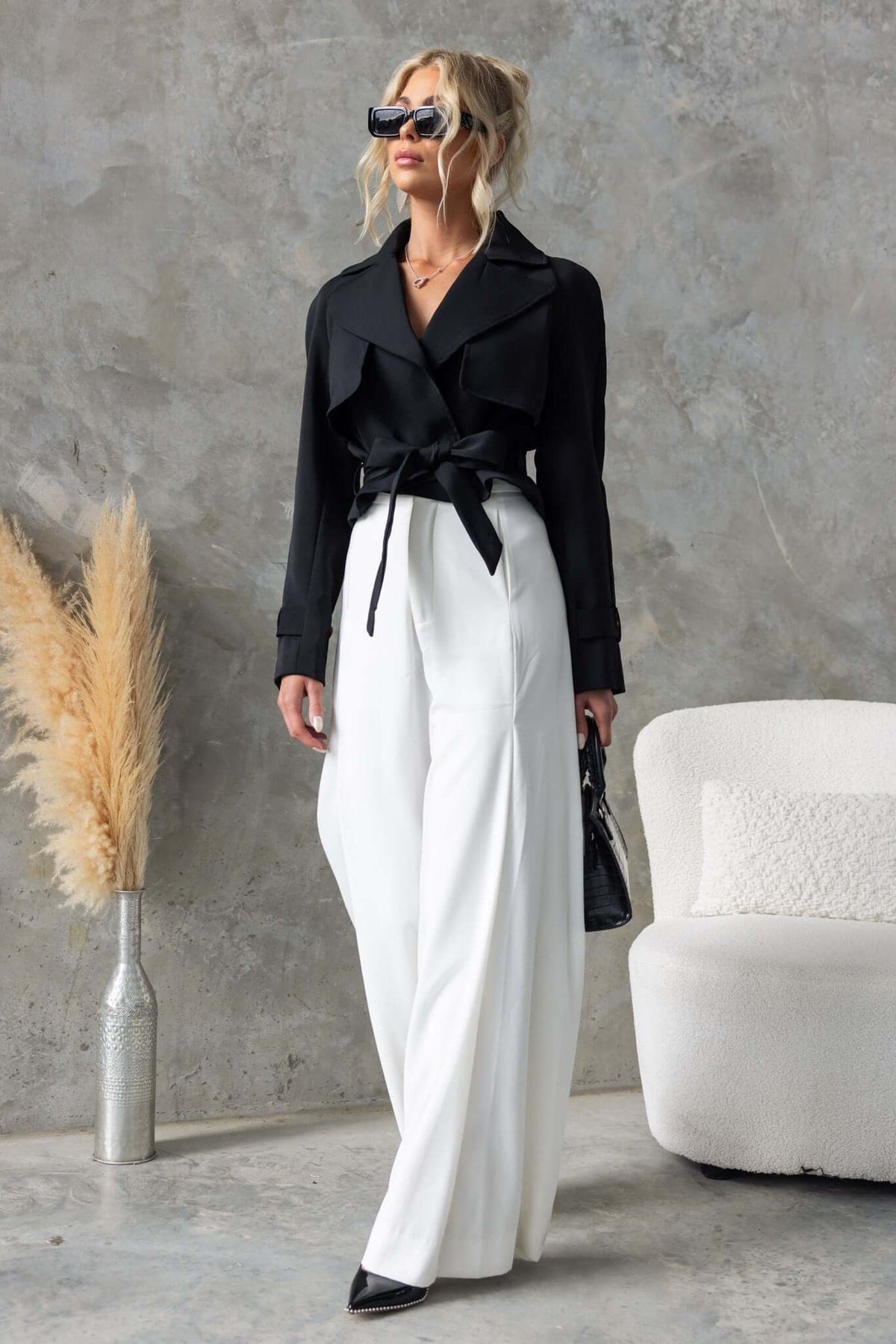 Model wearing the Ellis Coat in black with a removable tie, styled with white wide-leg pants and sunglasses.