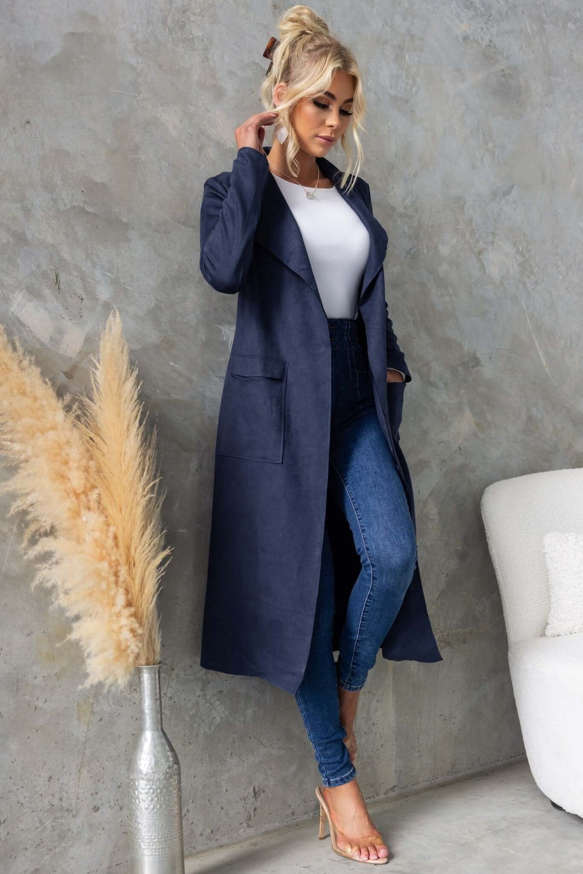 Model wearing a longline navy Hayley coat with draping lapel, styled with jeans in a modern interior.