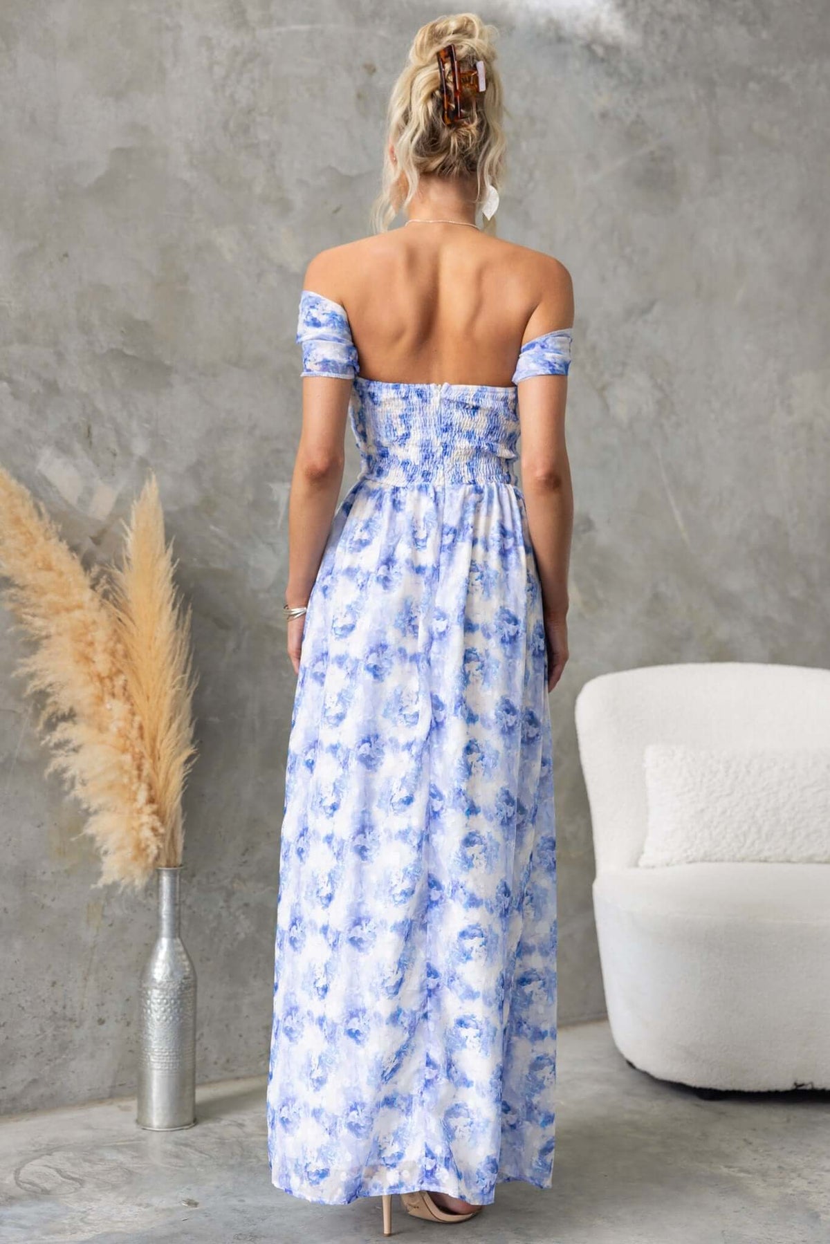 Back view of a model in a blue floral maxi dress with off-shoulder sleeves and asymmetrical hem, styled in a modern setting.