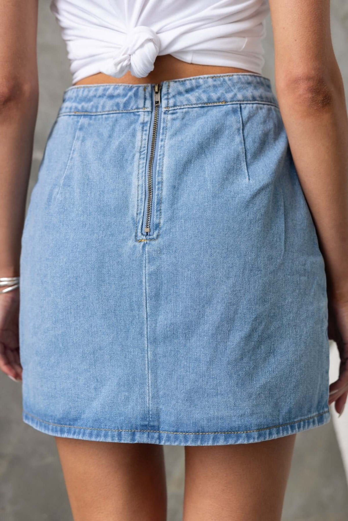 Back view of Lizzie Denim Skirt with a mid-high waist fit and zipper detail, styled with a knotted white top.