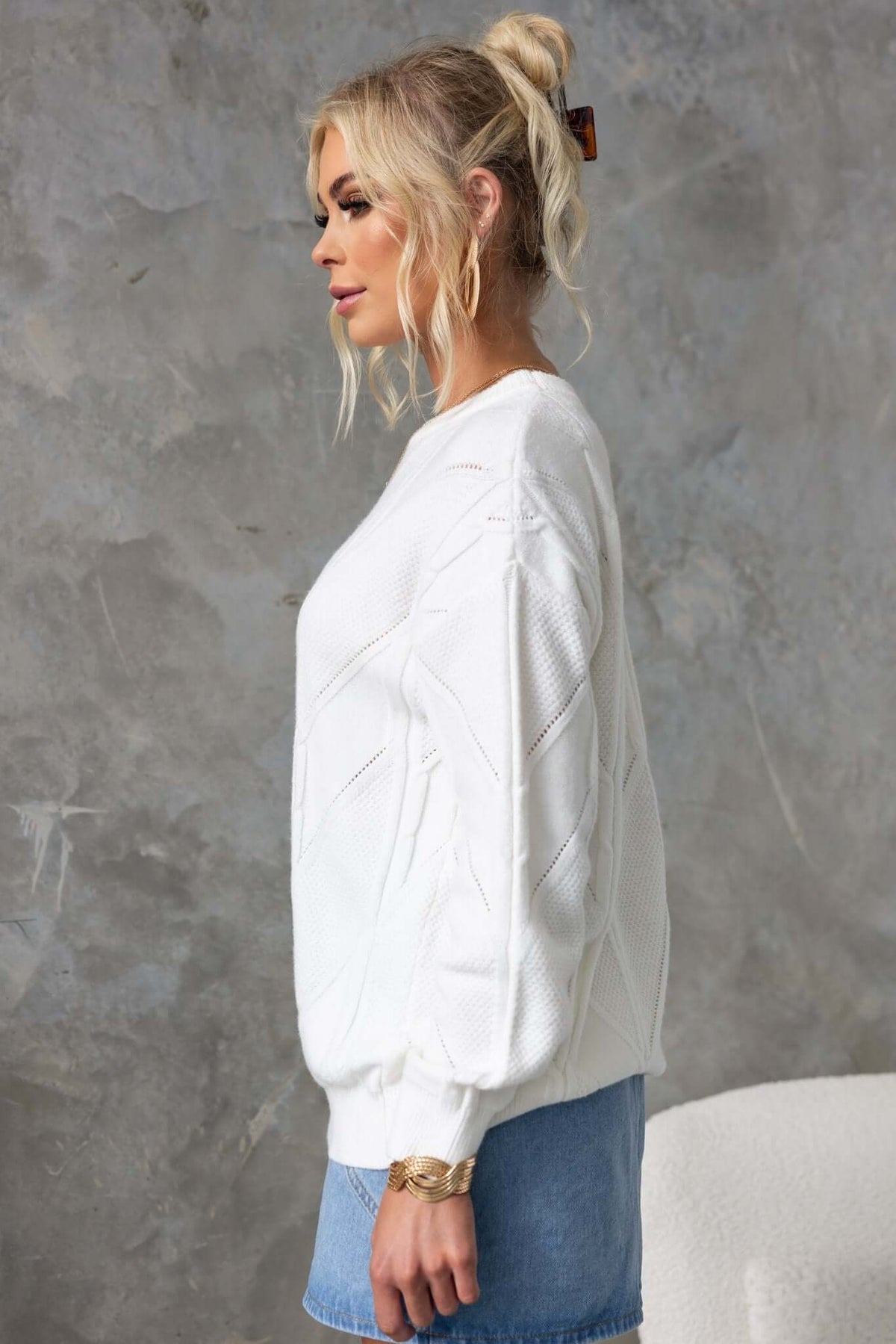 Side view of a woman in a white cable knit jumper, styled casually with a denim skirt and hair in a ponytail.