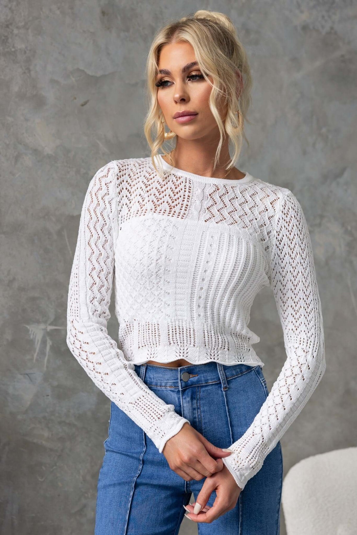 Model wearing a stylish white Perilla Top with long sleeves and intricate knit design, paired with blue jeans.