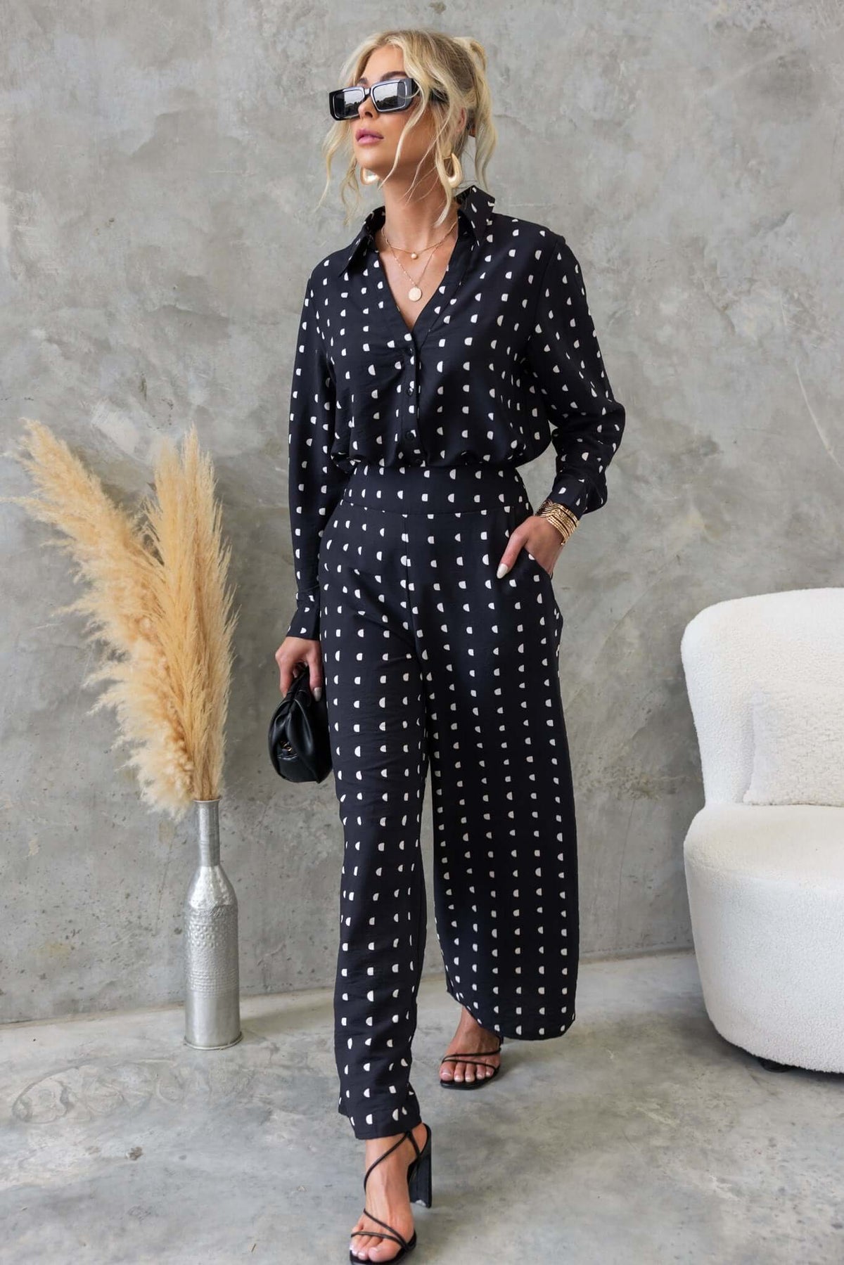 Woman in black Marion shirt with white polka dots, styled with sunglasses and heels, against a textured backdrop.