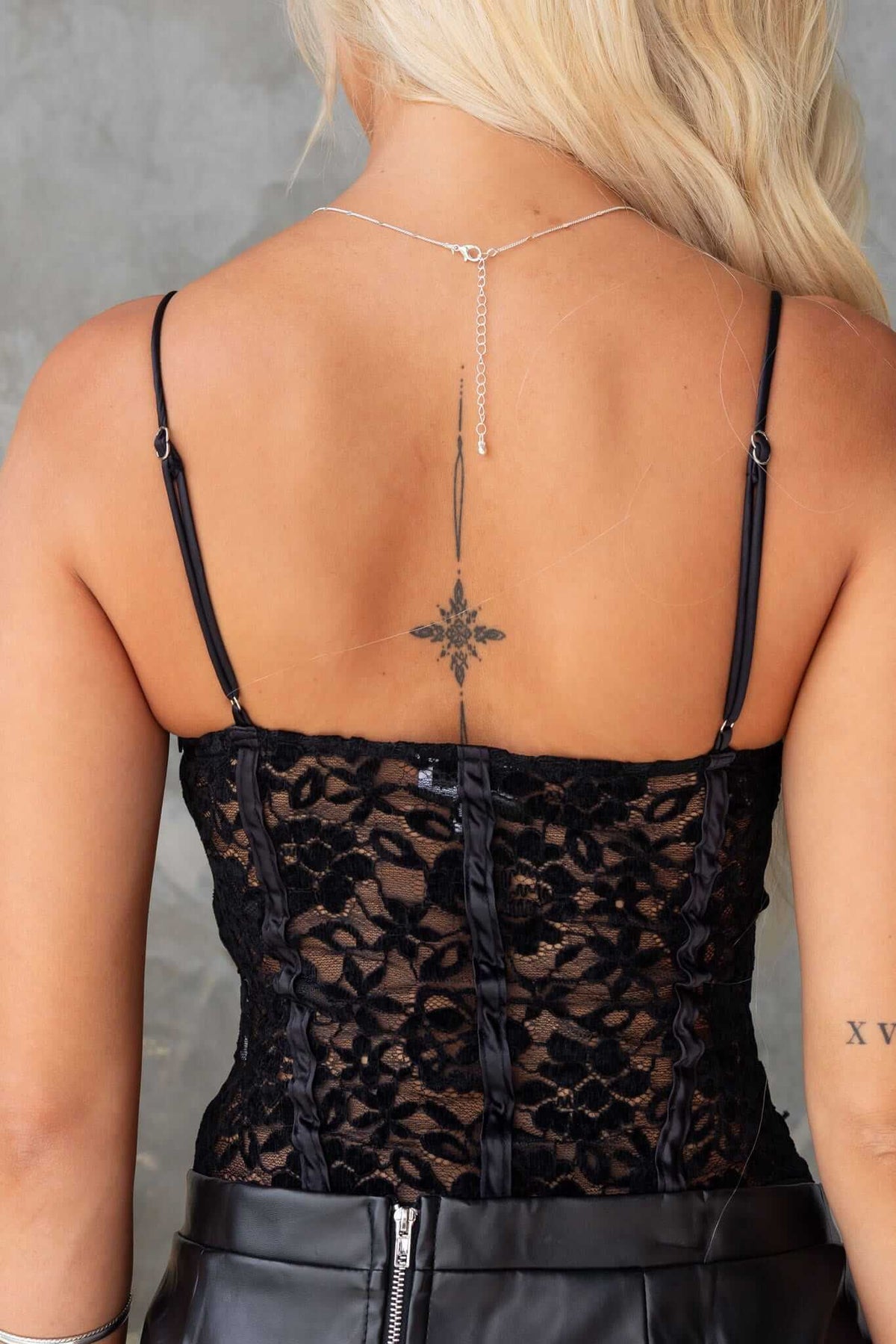 Back view of a woman wearing a black lace bodysuit with adjustable straps and a tattoo, showcasing intricate design and details.