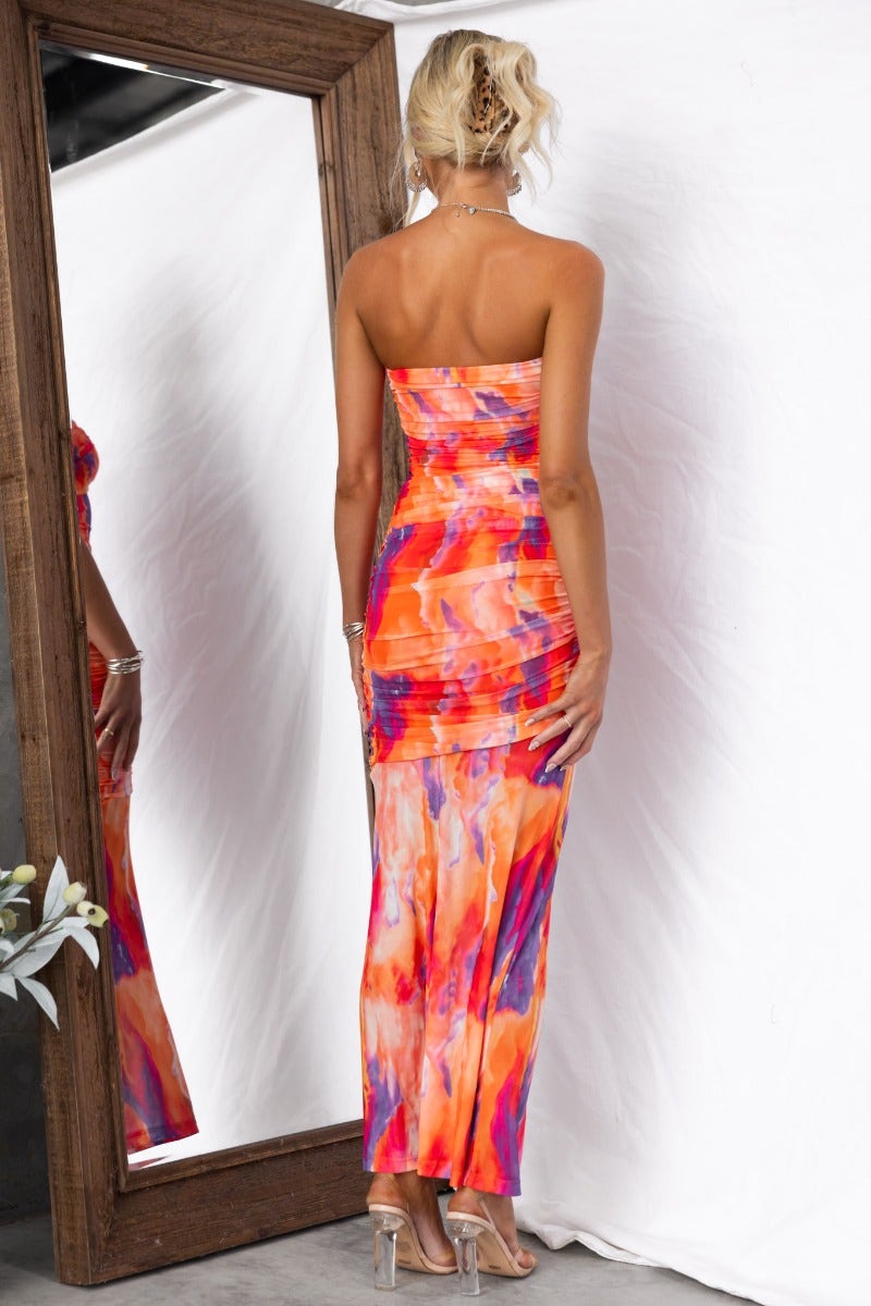 Model wearing an orange and purple strapless maxi dress, showcasing ruching and a mesh overlay, viewed from behind.