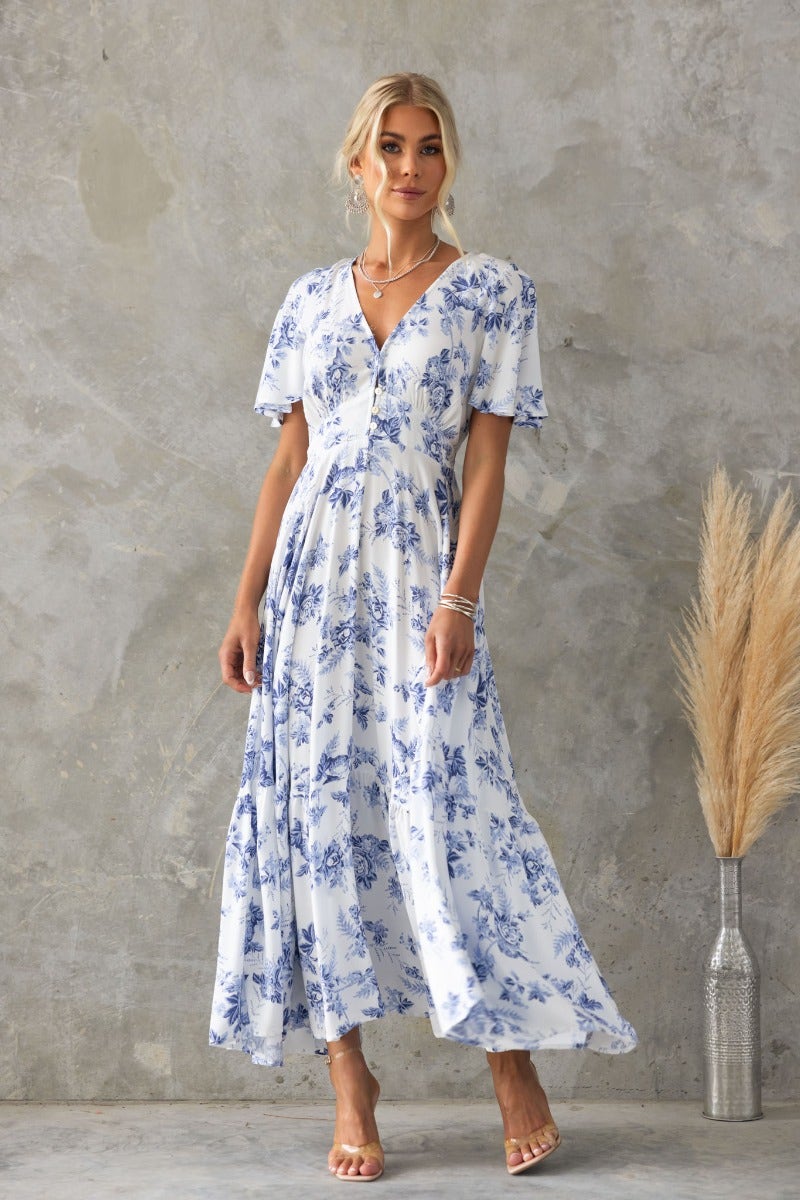 Nashville maxi dress with floral print, V-neckline, and short sleeves, styled with neutral accessories against textured backdrop.