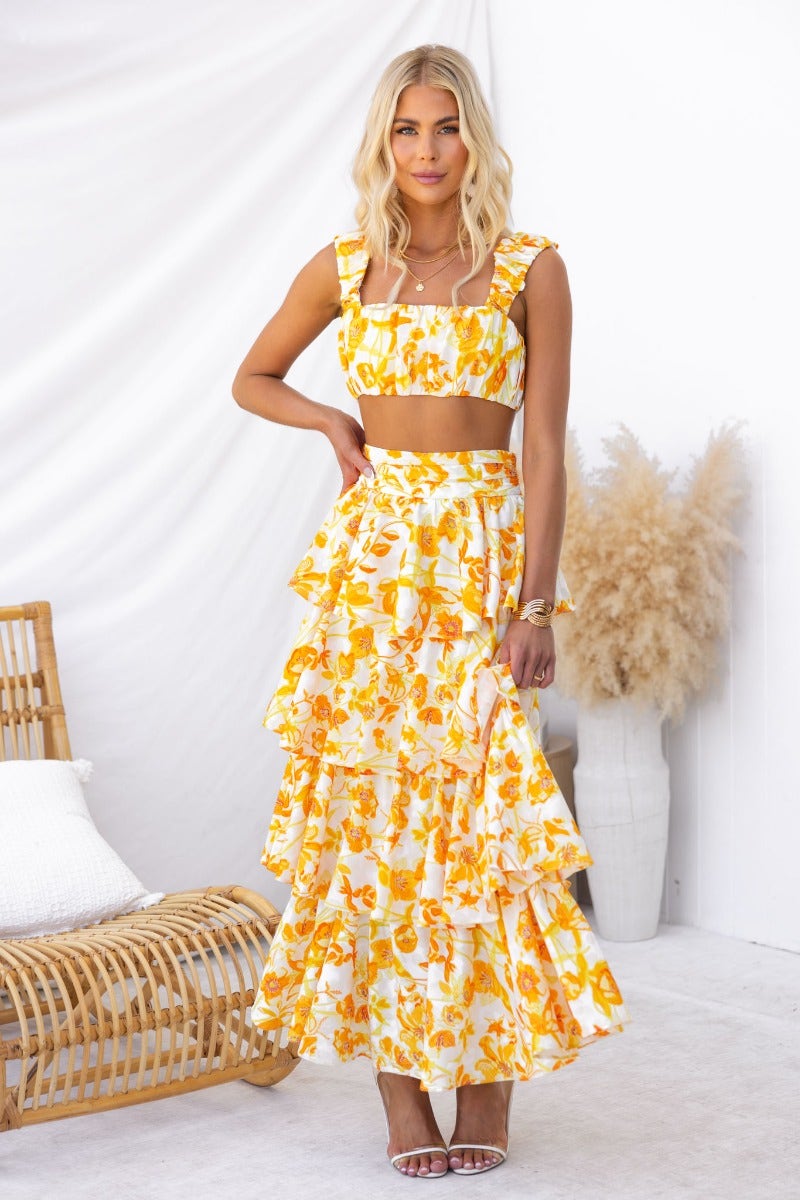 Model wearing floral print Illusion Maxi Skirt with ruffles and matching crop top, set in bright yellow tones.