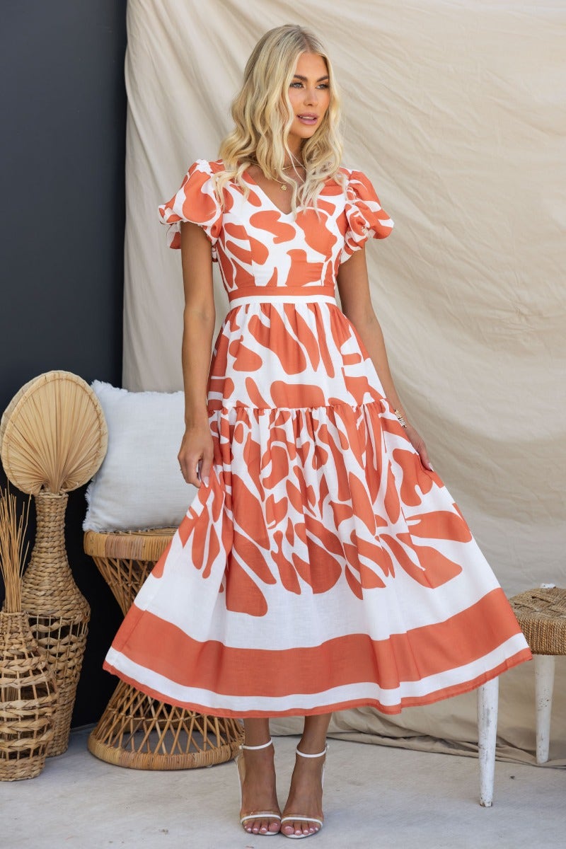 Model wearing Naela Midi Dress with abstract orange print and V-neckline, perfect for summer events.