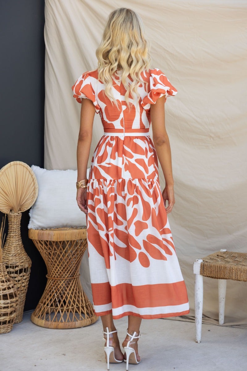 Back view of Naela Midi Dress featuring abstract print, V-neckline, and flutter sleeves in orange and white.