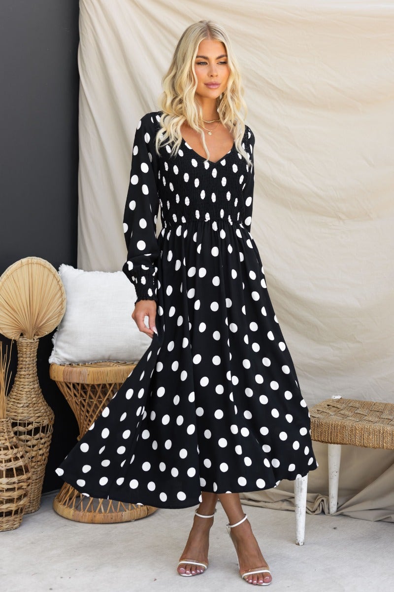 Dostan Midi Dress featuring a black polka dot design, V-neckline, and relaxed fit, styled with sandals.