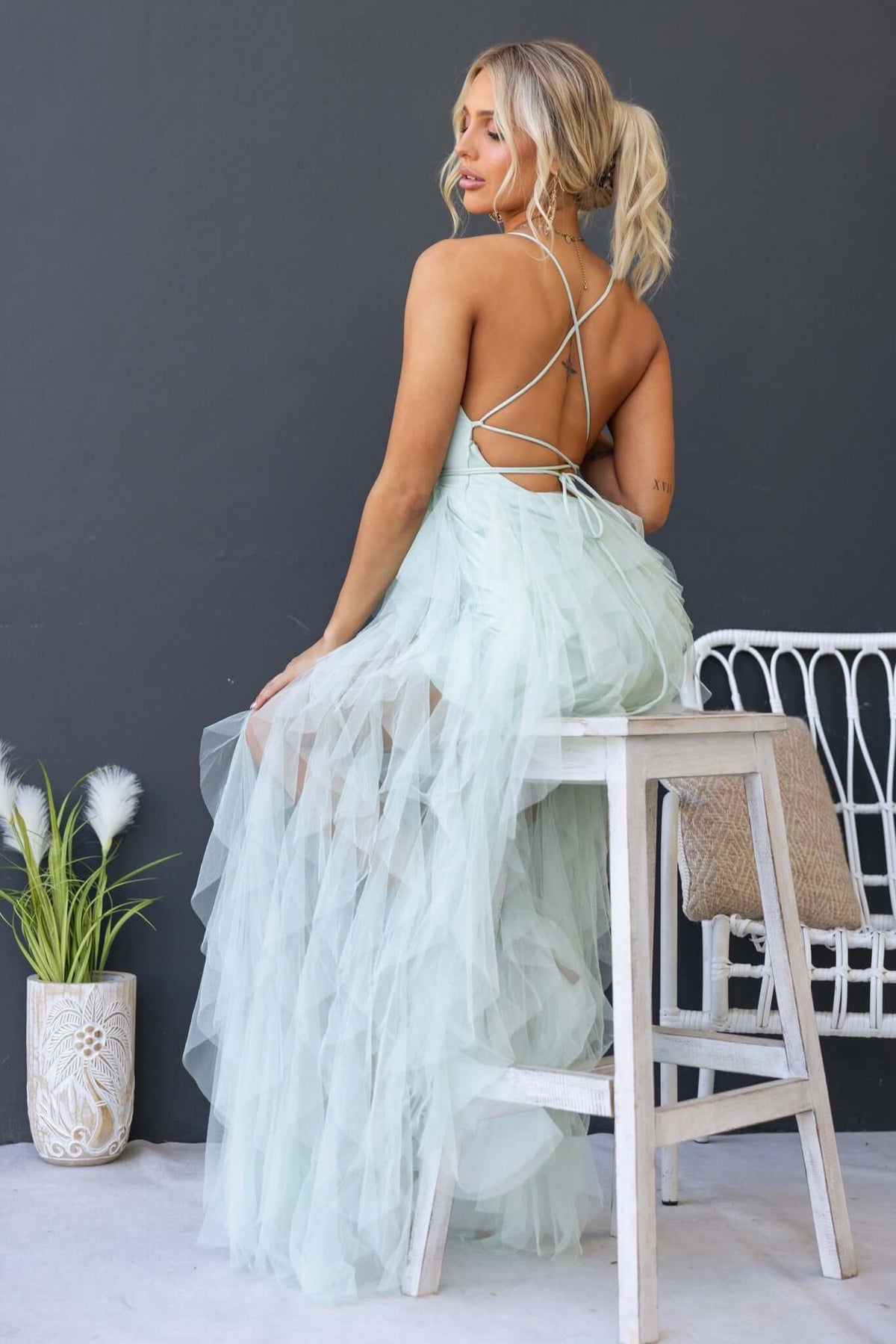 Woman in mint green Salinas Maxi Dress with lace-up back and tulle ruffle skirt, seated on a stool.