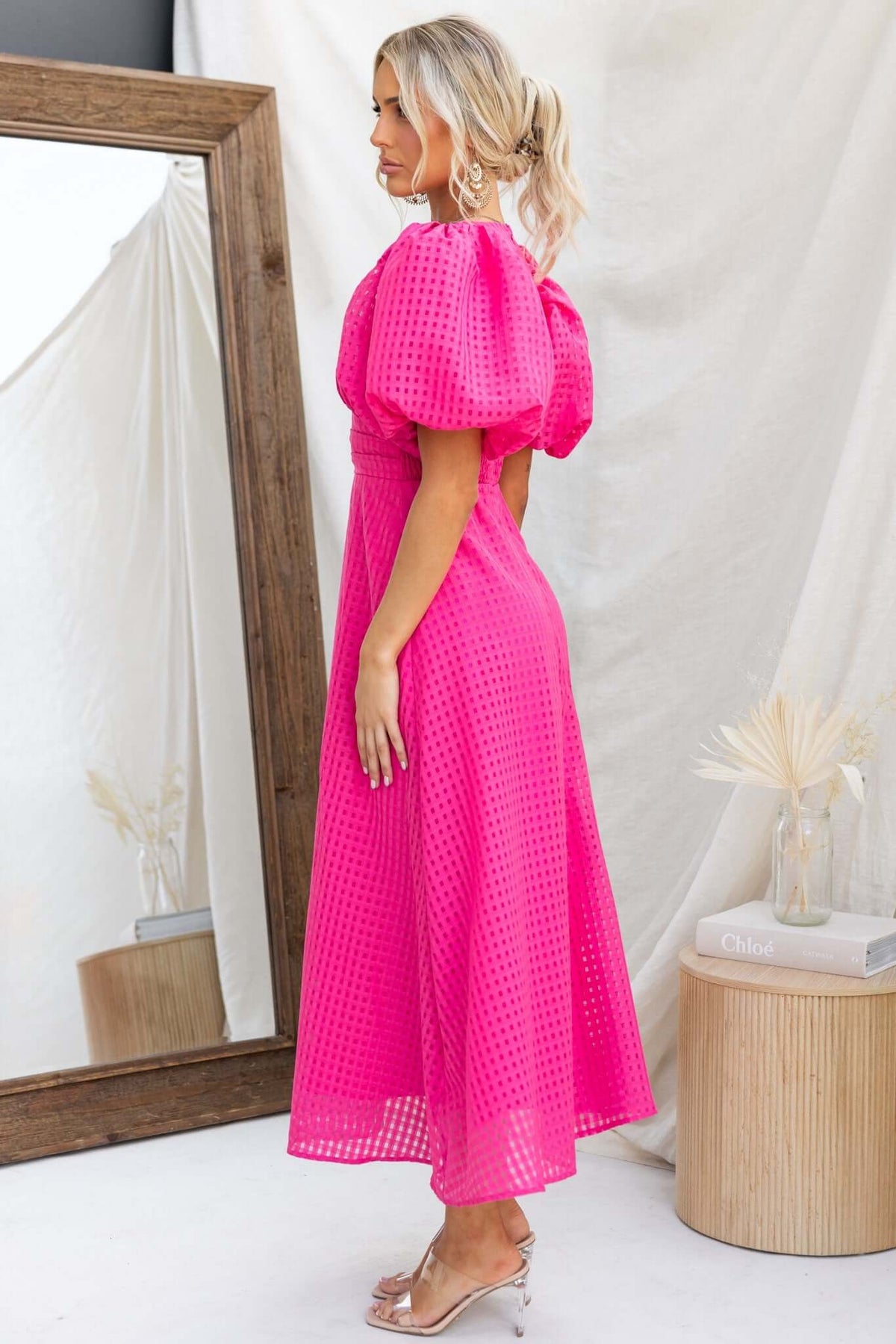 Model wearing a vibrant pink Nosita Midi Dress with puff sleeves and textured fabric, showcasing a stylish side view.