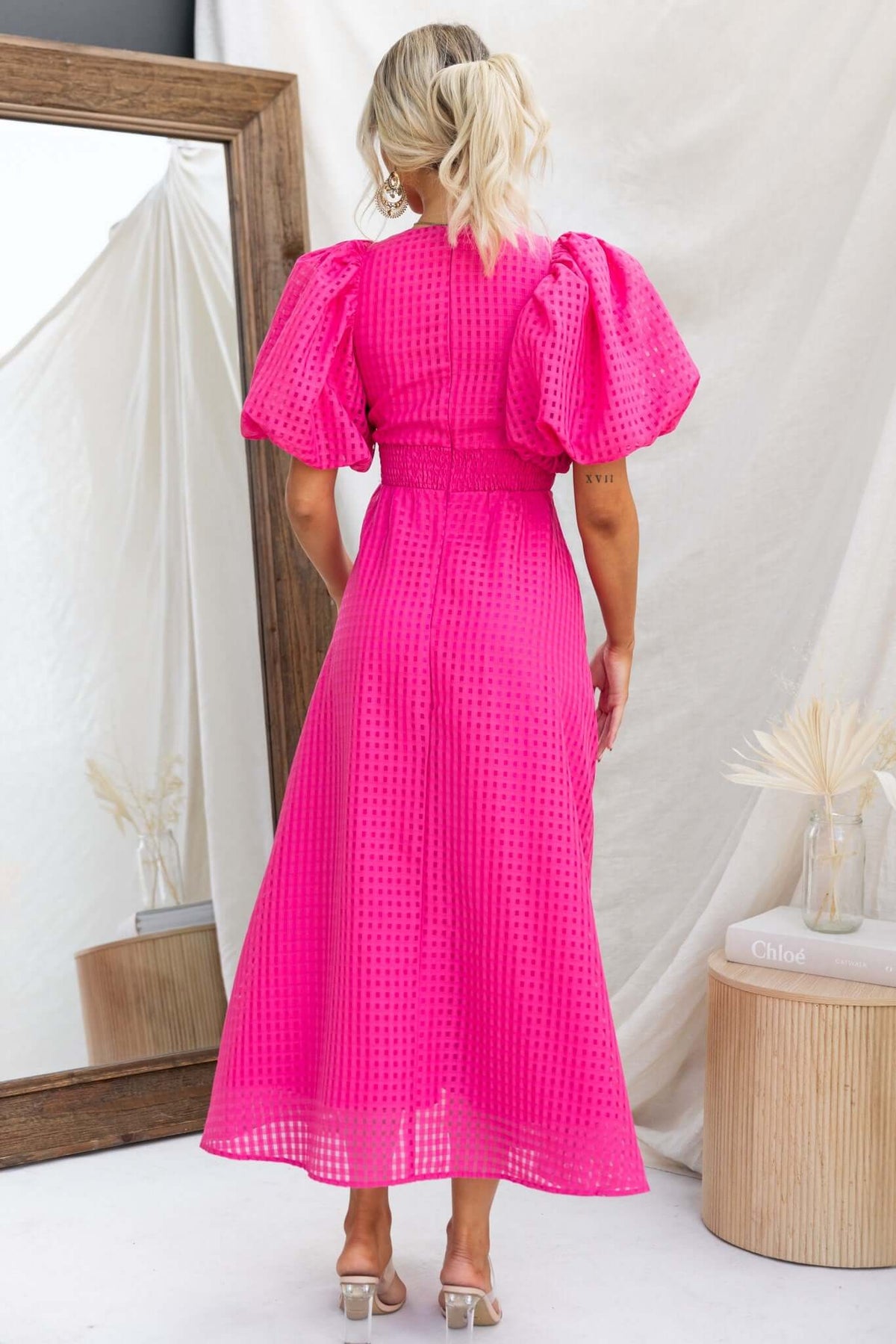 Back view of a woman in a bright pink Nosita midi dress with puff sleeves and textured fabric.