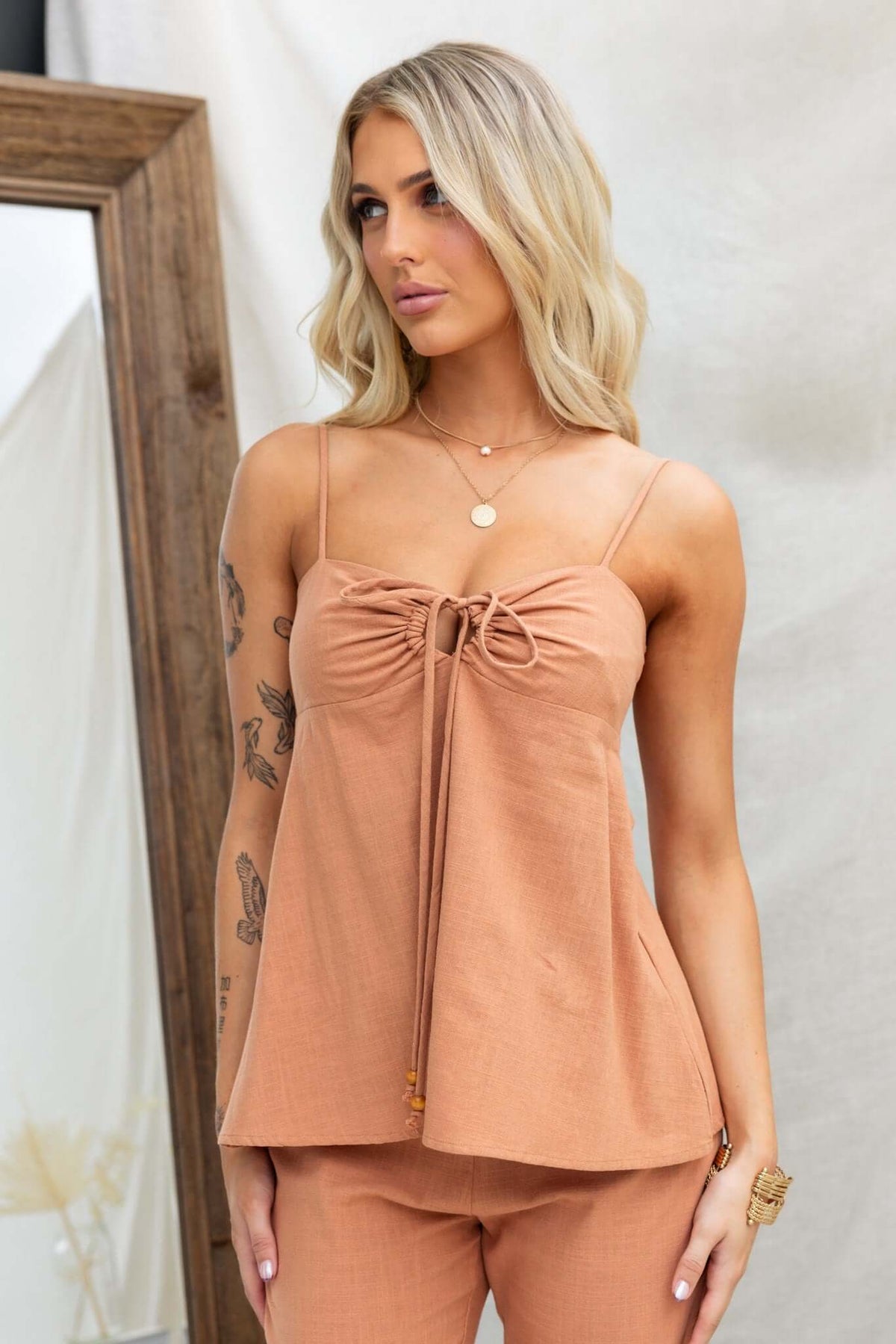 Model wearing the Stelline Top, a linen look adjustable strap top with a tie at the bust, in a warm peach color.