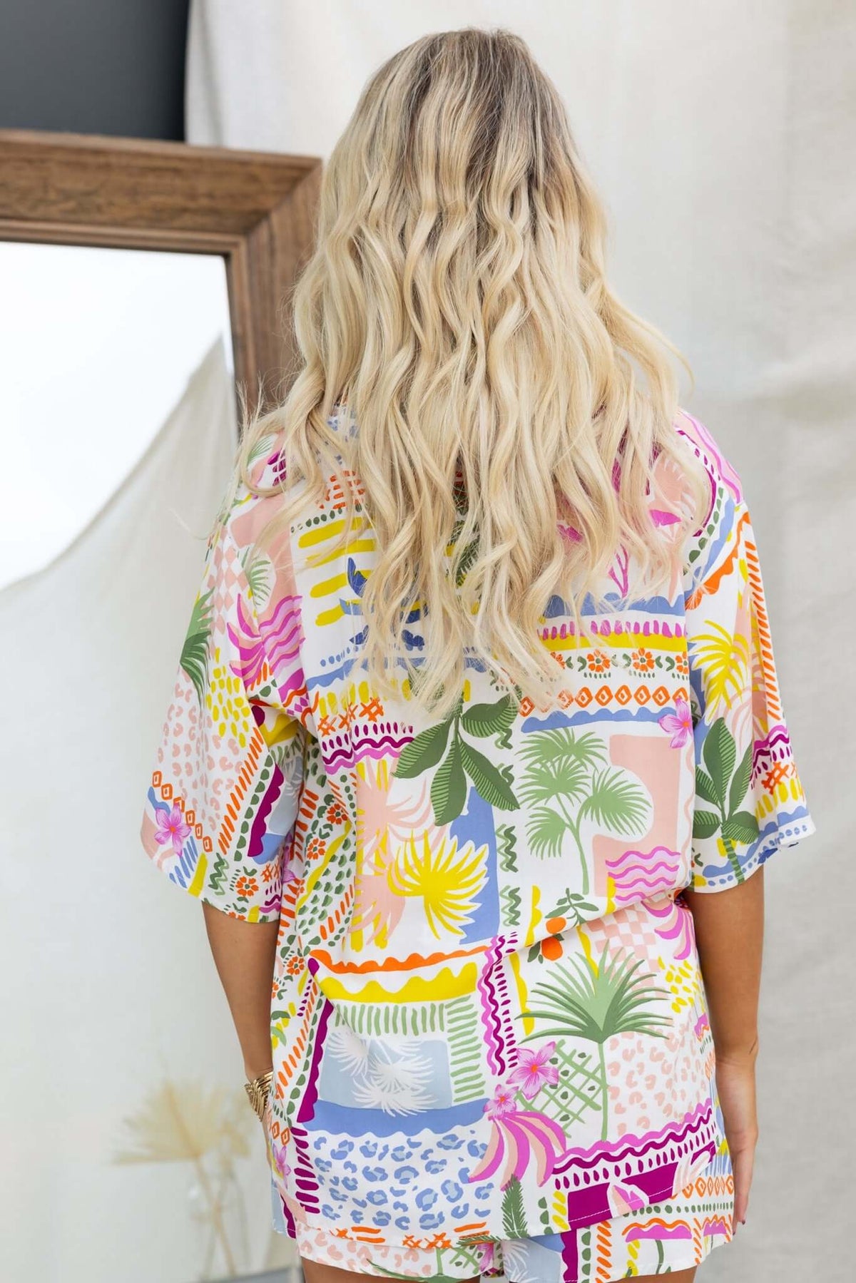 Back view of a woman wearing a colorful, patterned shirt with tropical designs, showcasing vibrant colors and relaxed fit.