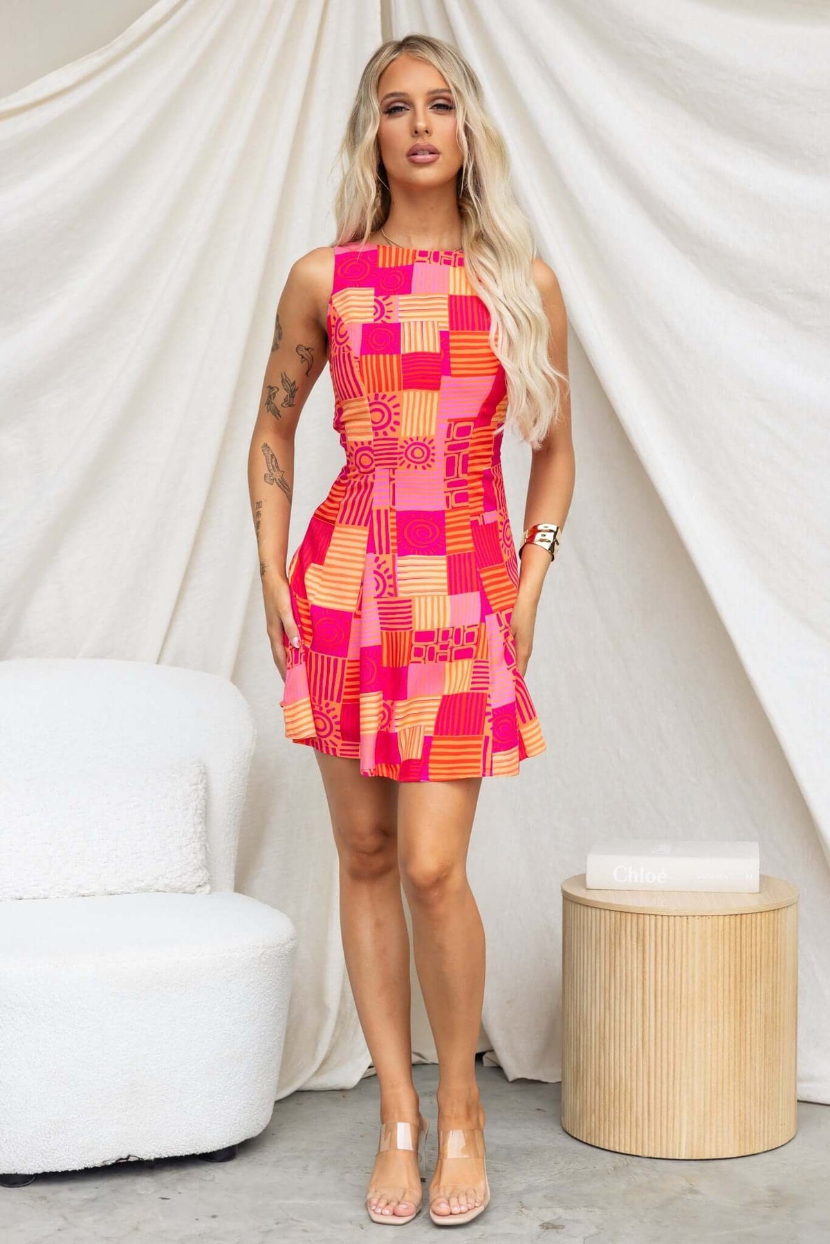 Model wearing a vibrant pink and orange patterned sleeveless dress, standing next to a minimalist round table.
