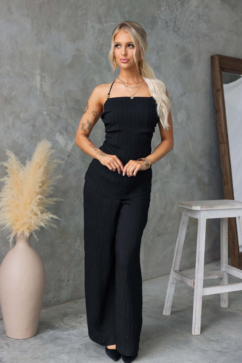 Stylish woman in black Kayce Set, featuring tie neckline and mid-high waist pants, posed elegantly against a modern backdrop.