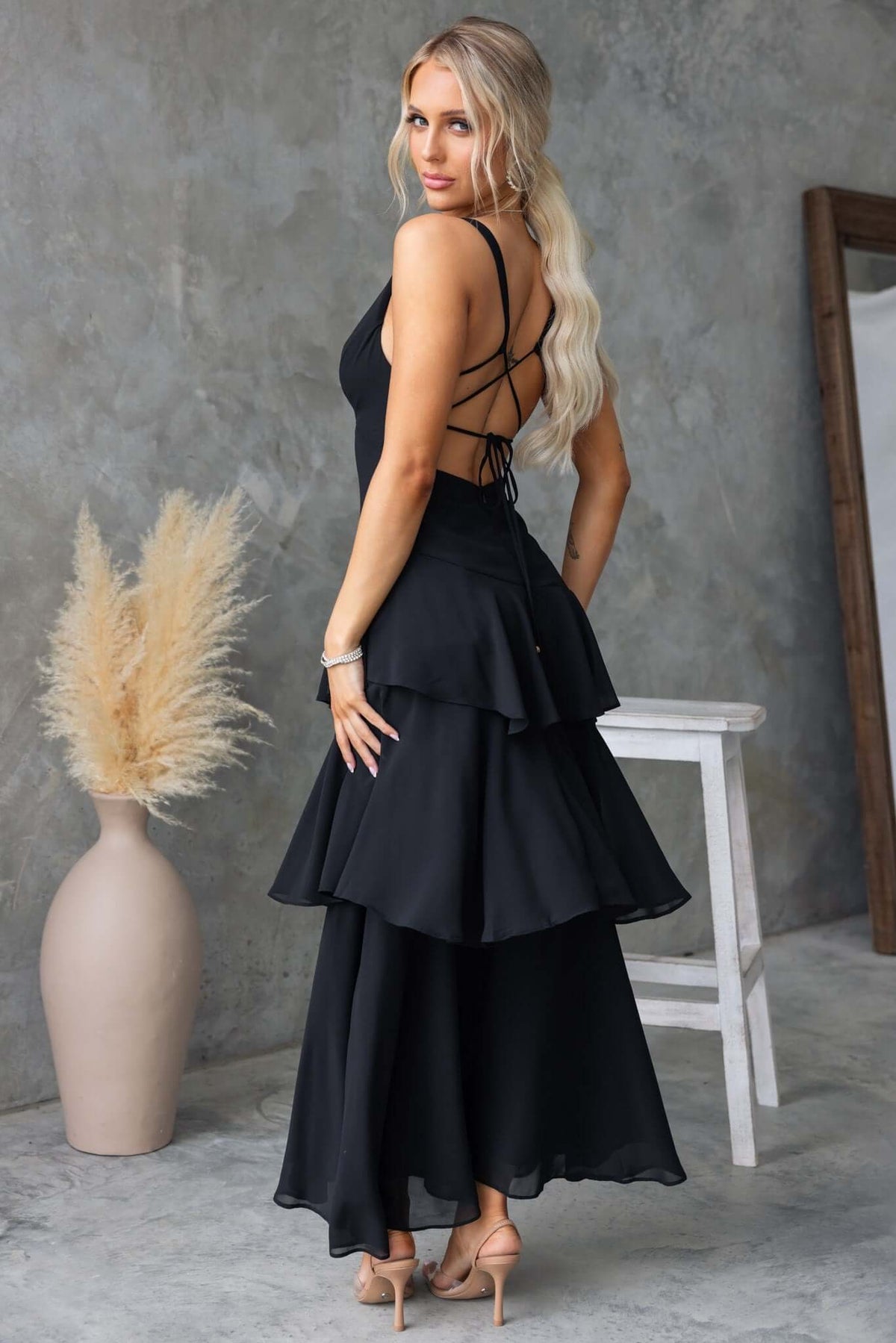 Back view of a woman in a black maxi dress with multi-tier ruffle hem and tie detail at the back.