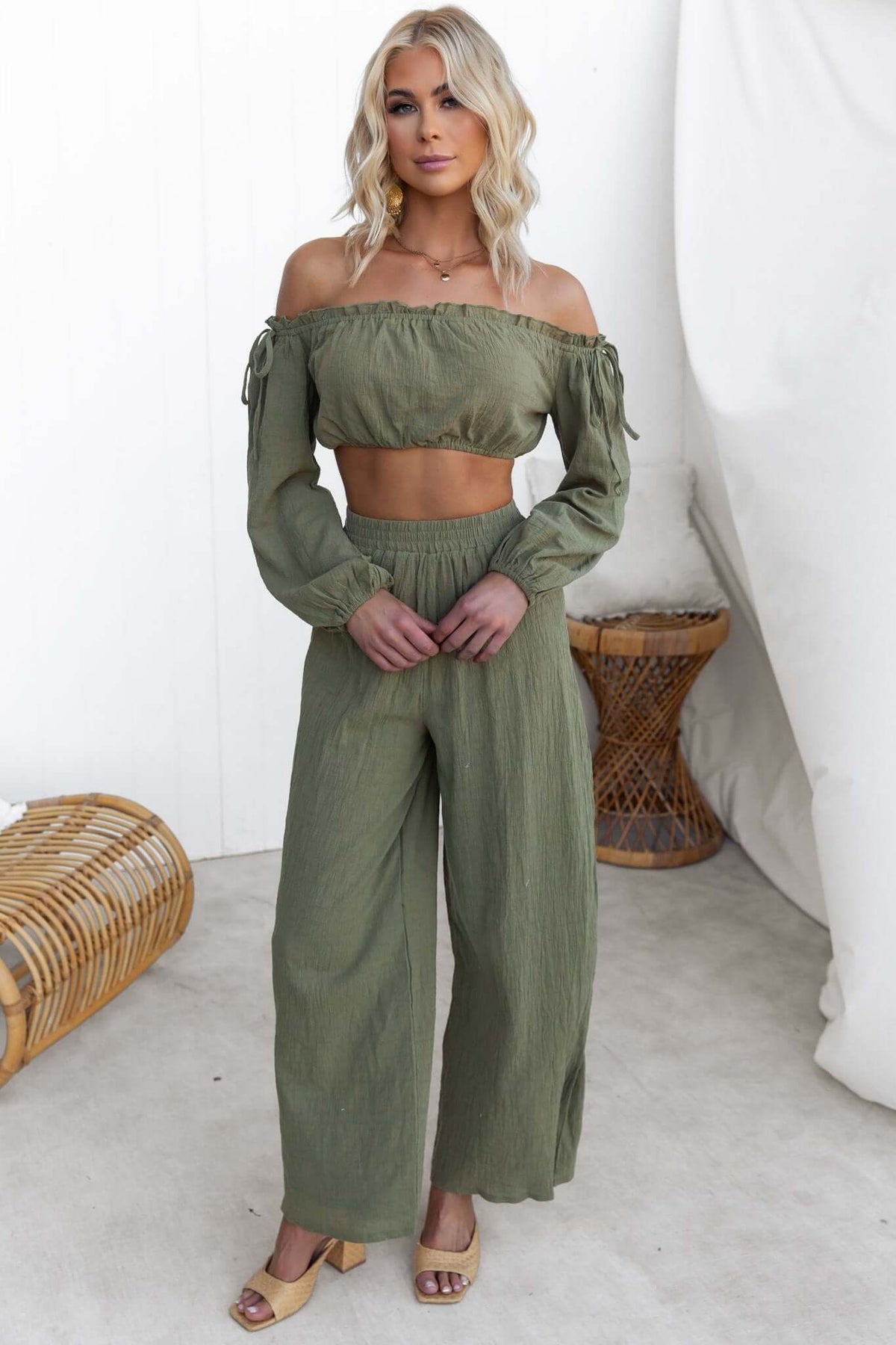 Model wearing an olive green off-shoulder Gravina Top with tie details, paired with wide-legged pants in a stylish setting.