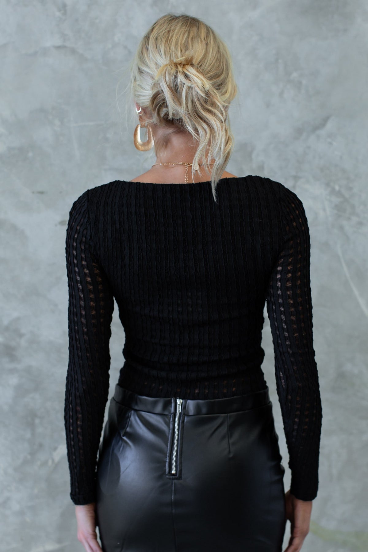 Back view of a woman in a black textured top with a boat neckline and long sleeves, paired with a sleek black skirt.