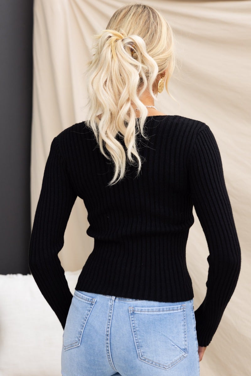 Back view of a woman in a black ribbed knit top with long sleeves and V-neck, paired with light blue jeans.