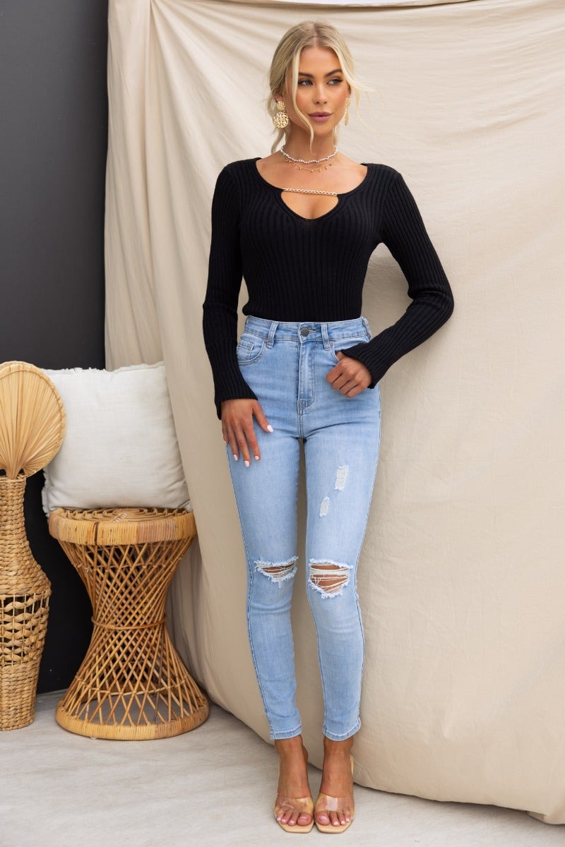 Model wearing the Ferla Top, a black ribbed knit V-neck with chain detail, paired with light blue ripped jeans.