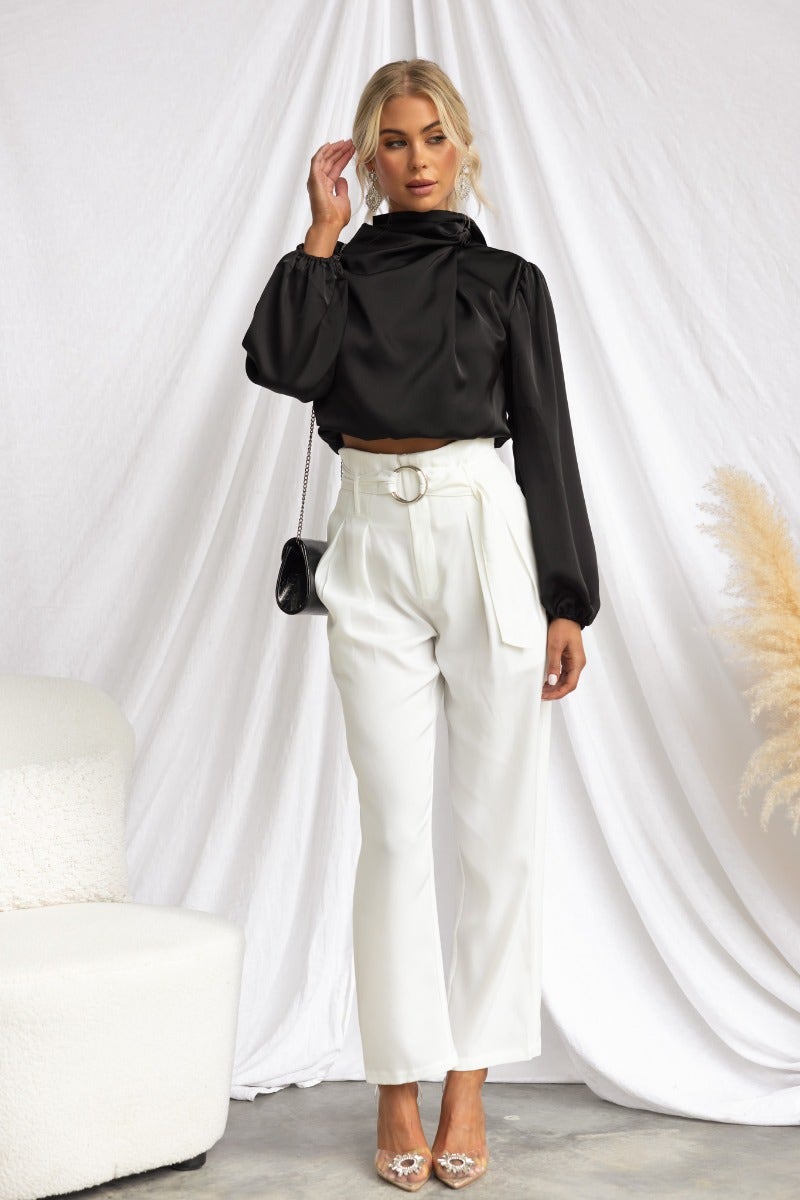 Model wearing a black satin Xali top with high neckline, paired with white pants for a stylish look.