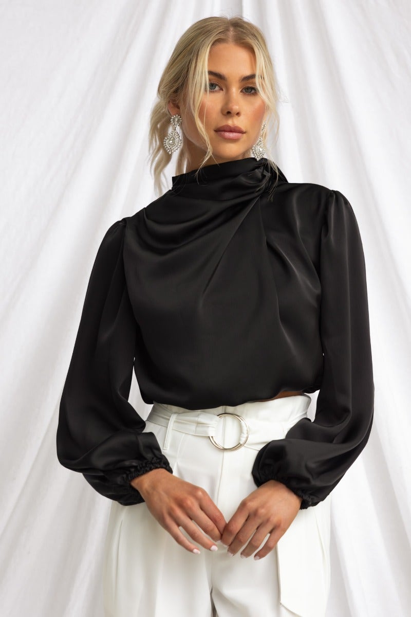 Black satin Xali Top with high neckline and elastic sleeve cuffs, styled with white pants.