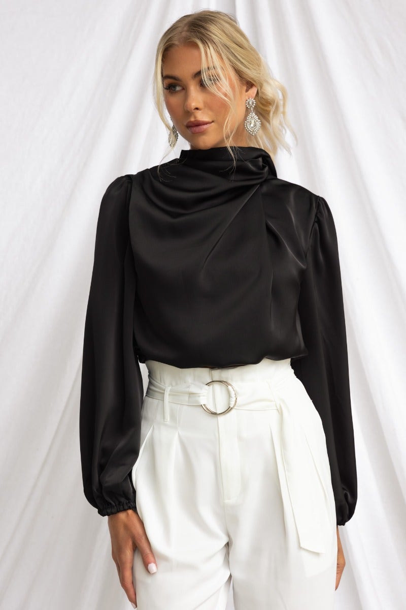 Black satin Xali Top with high neckline and elastic cuffs, styled with white pants and earrings.