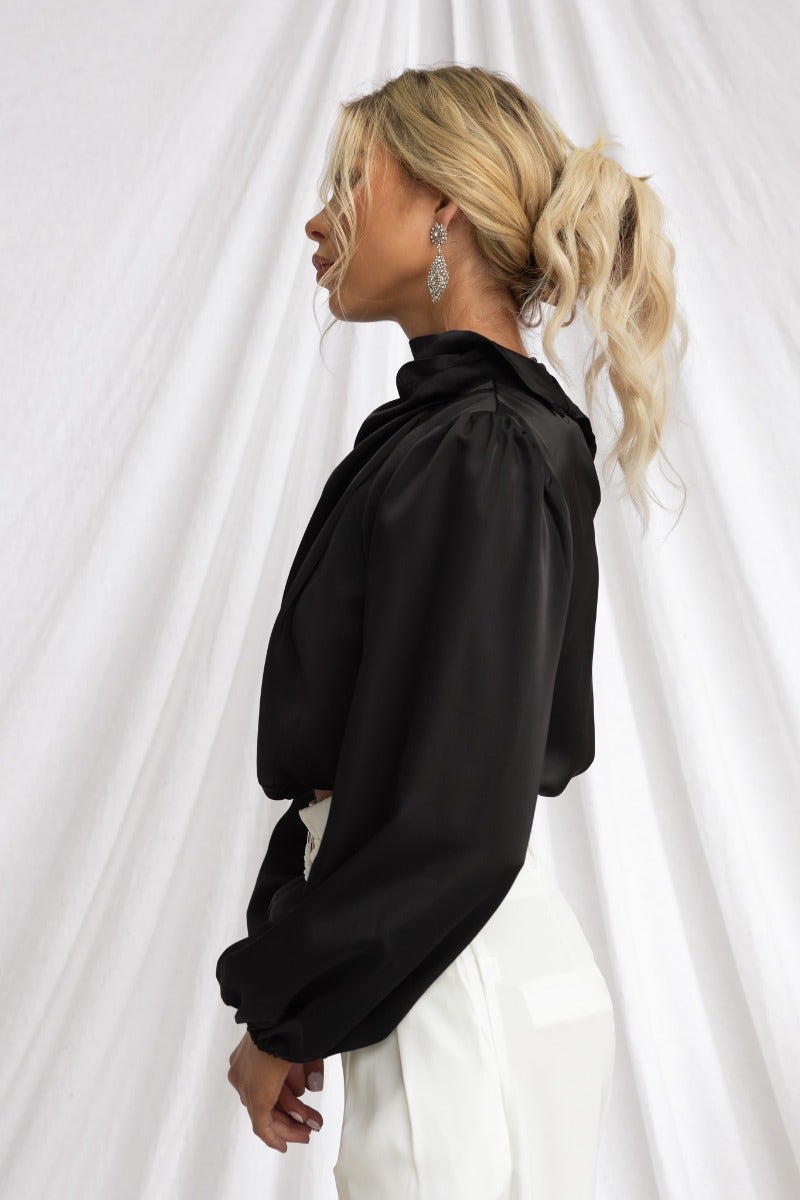 Profile view of a woman wearing a black satin high neckline top with balloon sleeves.