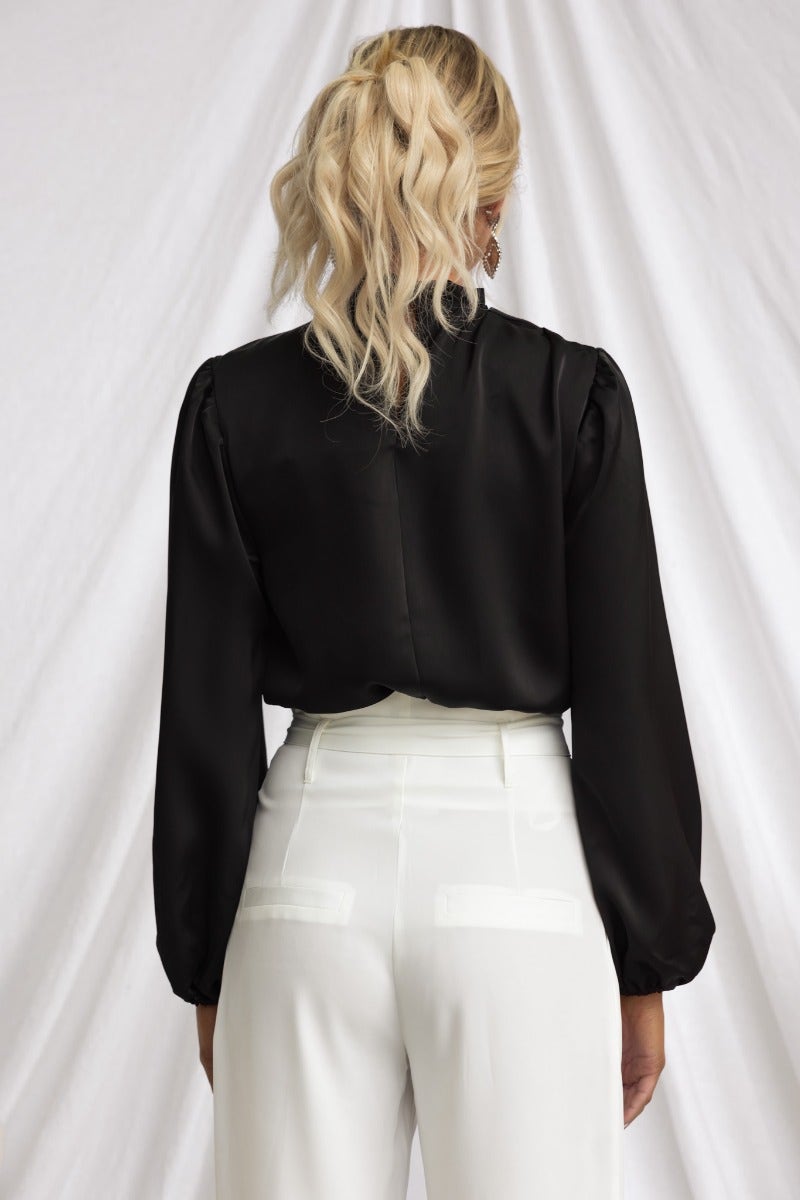 Black satin top with high neckline and cuffed sleeves, styled with white pants, showcasing the back view.