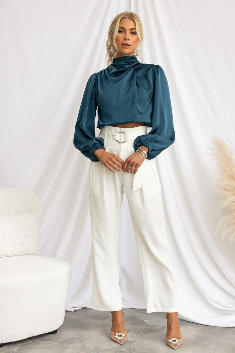 Model wearing a teal satin high-neck top with elastic cuffs, paired with white tailored pants in a stylish setting.