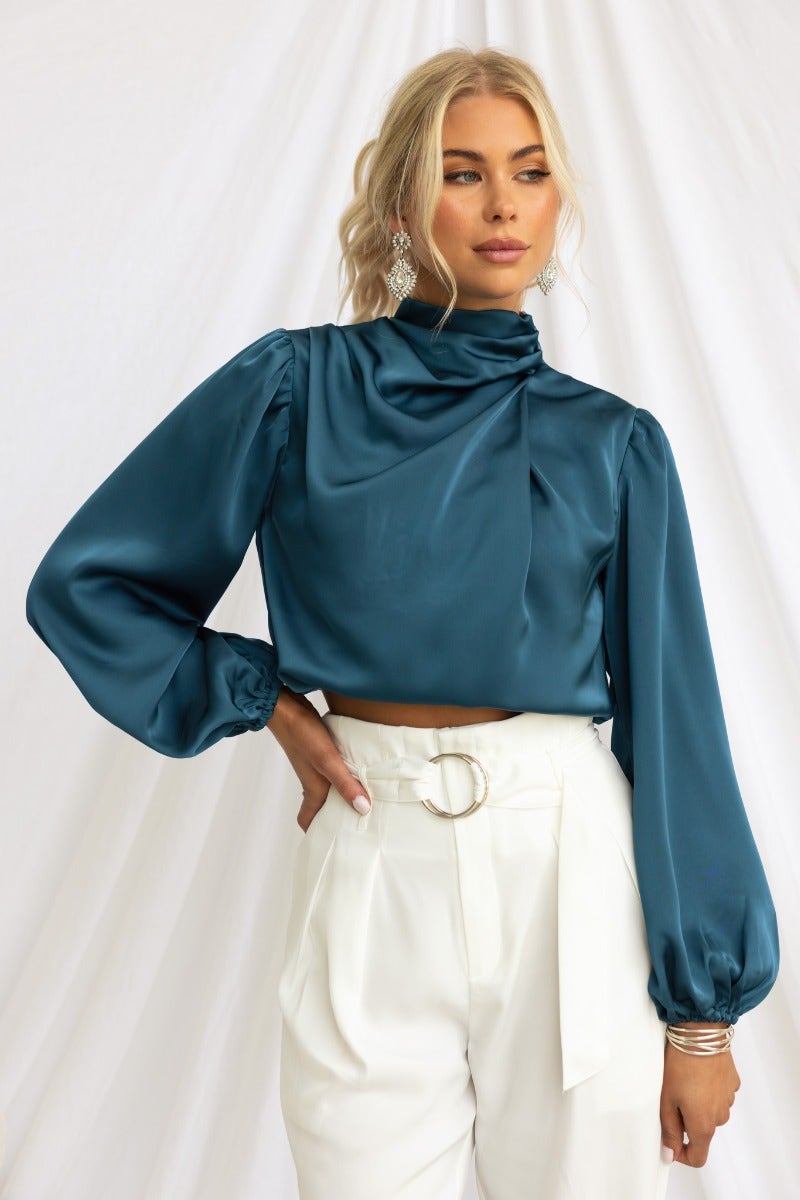 Xali Top in teal satin with high neckline, balloon sleeves, styled with white high-waisted pants. Perfect for elegant occasions.