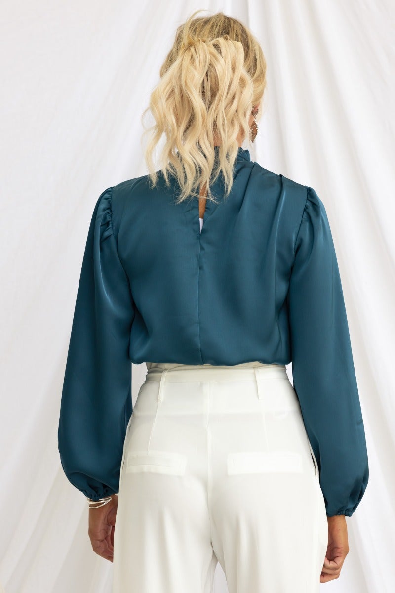 Back view of teal satin Xali Top with high neckline, puffed sleeves, and buttons at the neck, styled with white trousers.