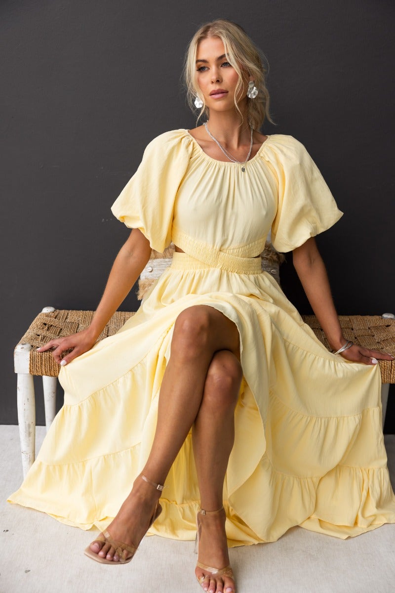 Model wearing a Posh Maxi Dress with an elastic off-shoulder neckline and cut-out at waist, sitting elegantly on a bench.