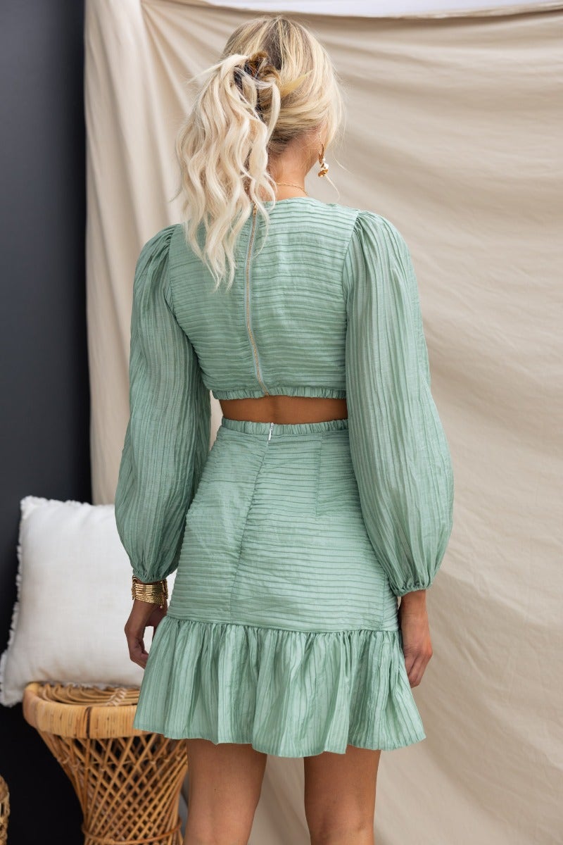 Back view of Tovia Dress with textured green material, puff sleeves, and ruffled hem, showcasing stylish cut-out at waist.