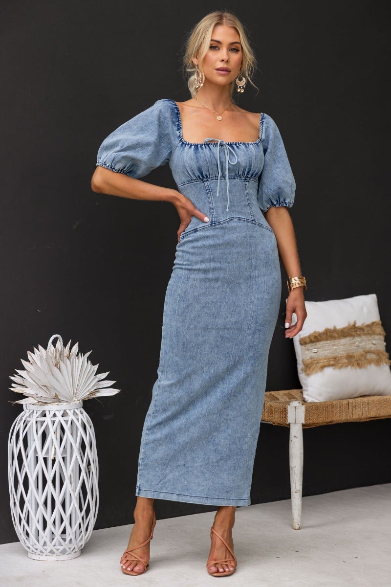 Model wearing Irving Denim Maxi Dress with puff sleeves, tie detail at bust, and a back split, set against a stylish backdrop.