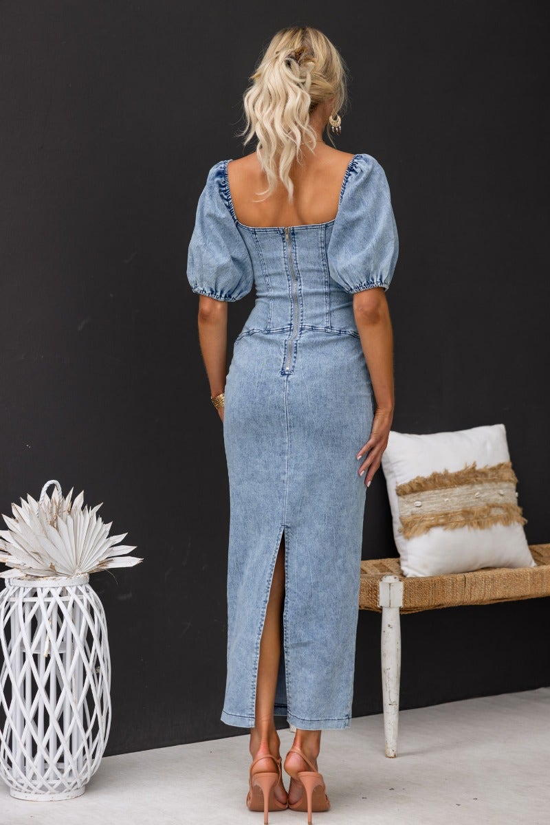 Back view of Irving Denim Maxi Dress with puff sleeves, zip detail, and split, styled with accessories in a chic setting.