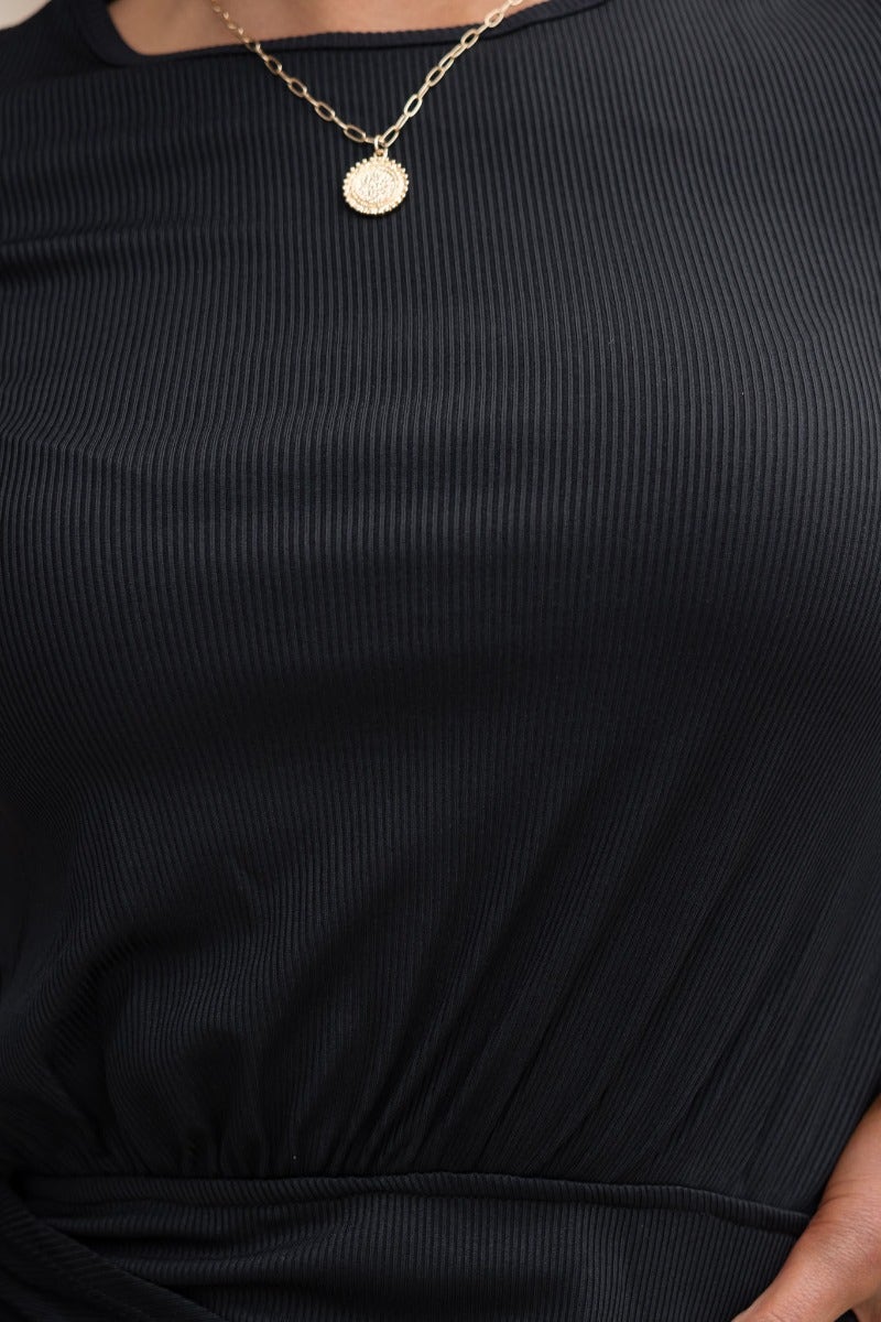 Close-up of black ribbed Devia dress showcasing elastic waistband and twist detail, paired with a delicate gold necklace.