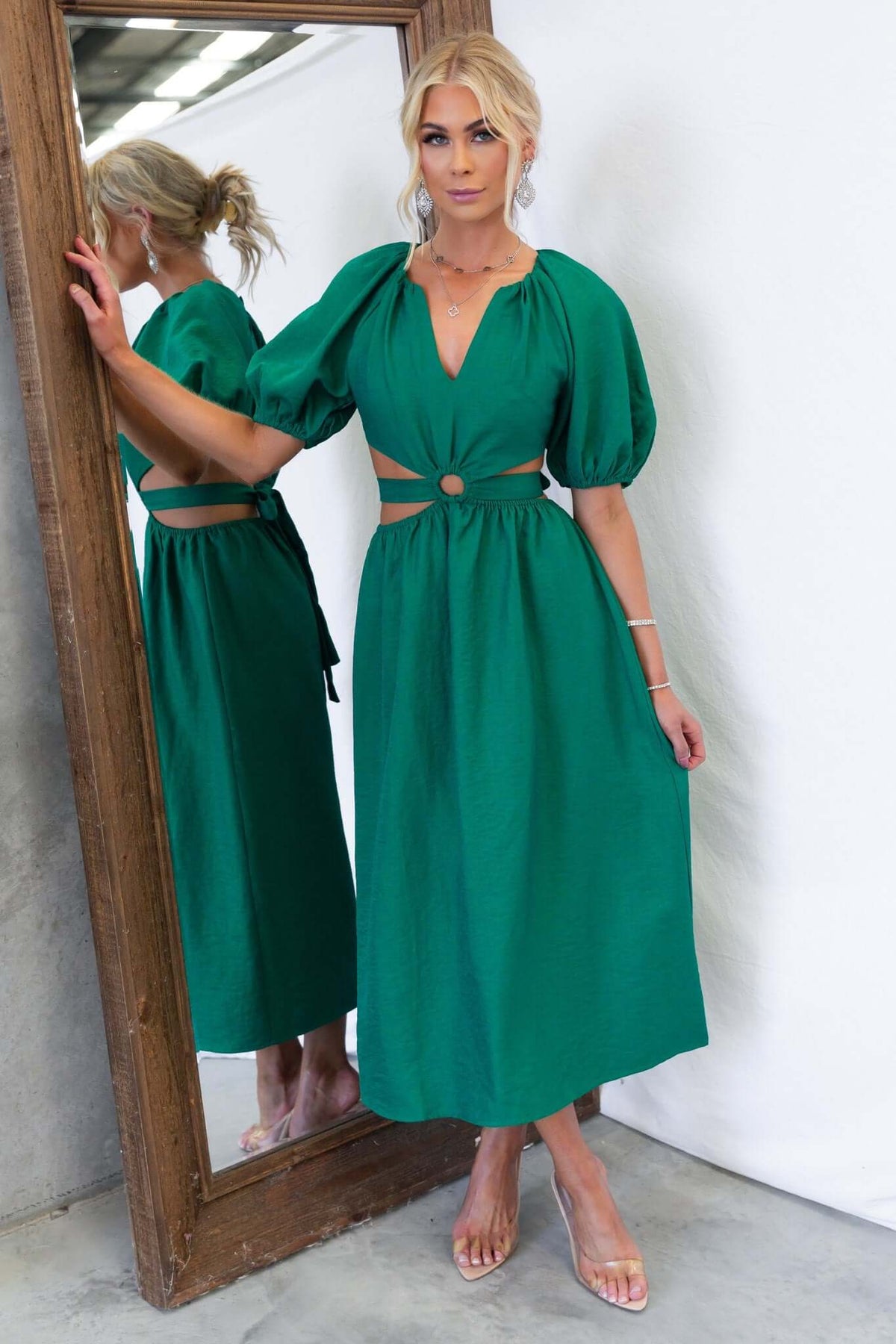 Model wearing a green midi dress with a V-neckline, puff sleeves, and cut-out waist, posed by a mirror.