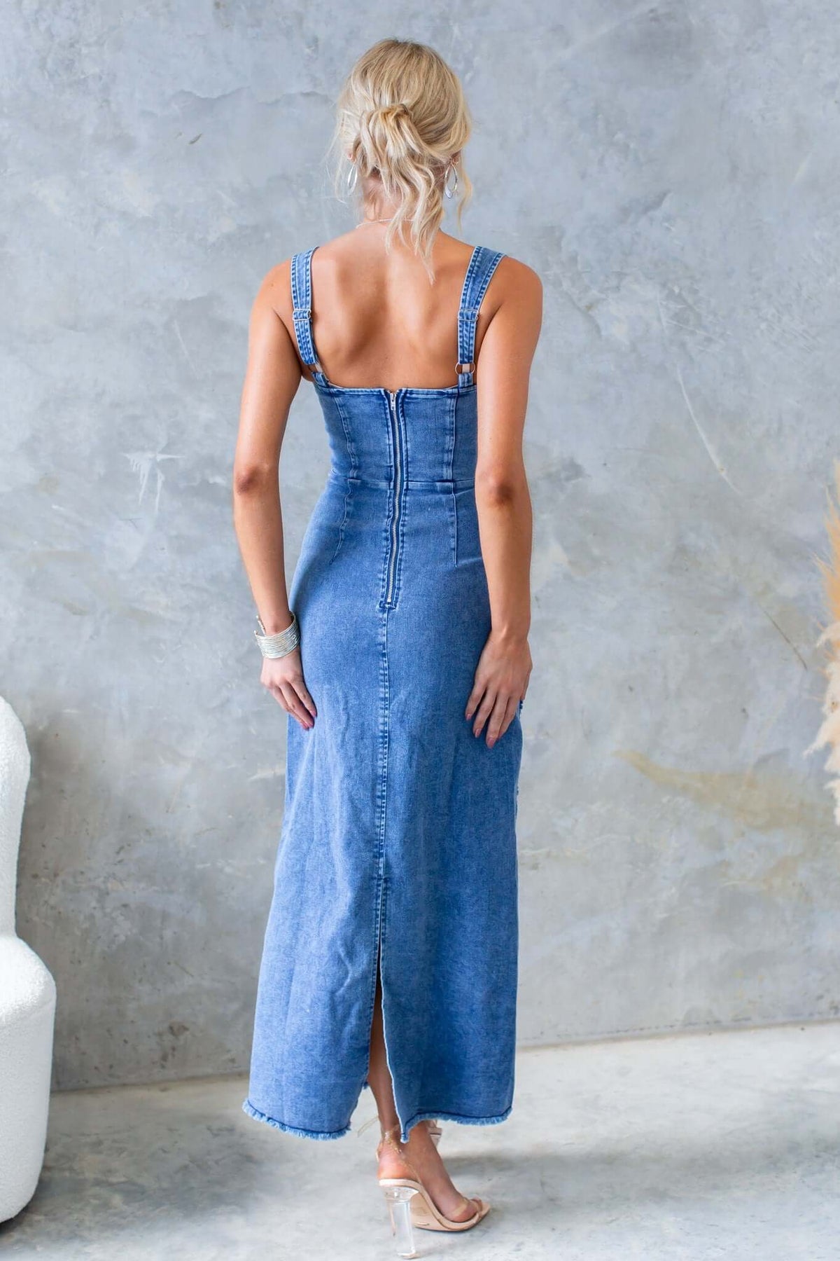 Back view of a woman wearing a denim maxi dress with adjustable straps and a zip closure, featuring a back split.