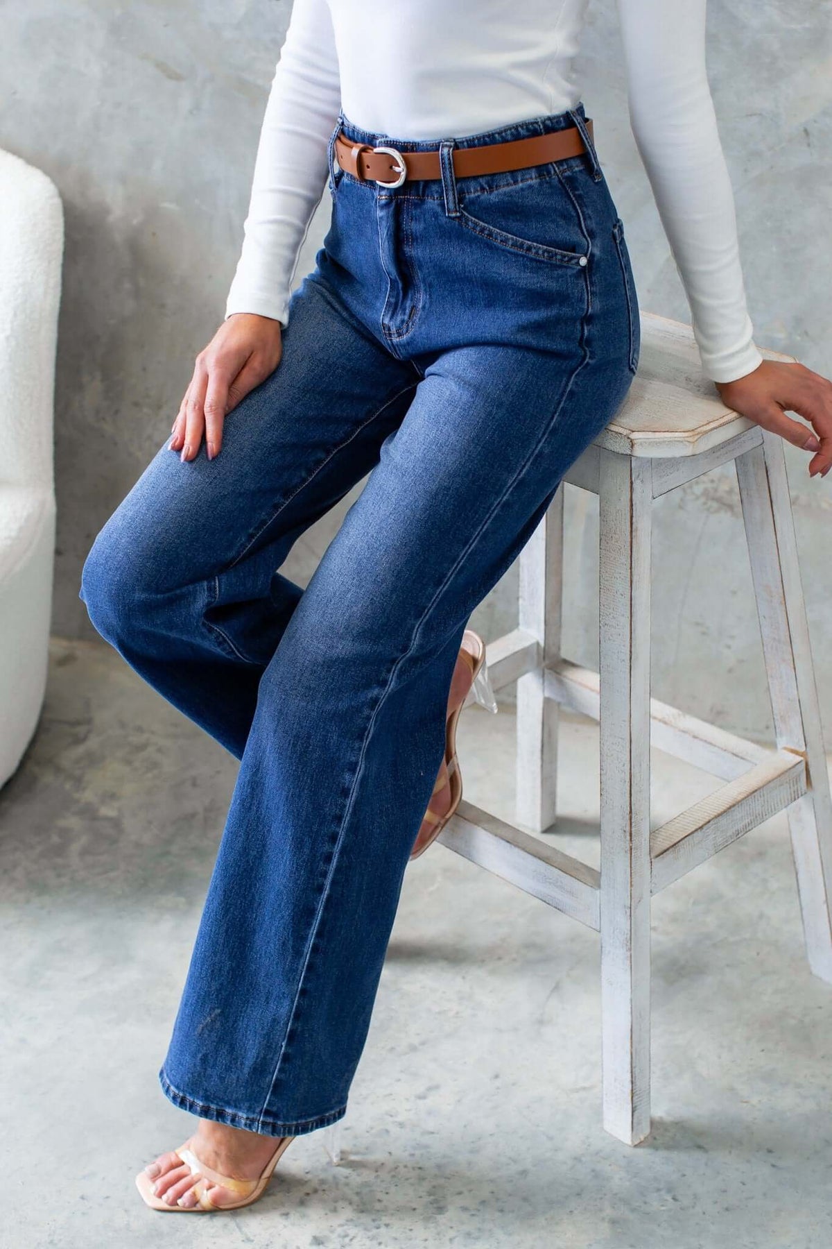 Model wearing Wallen Jeans with mid-high waist, wide legs, and a stylish belt, seated on a stool against a neutral backdrop.