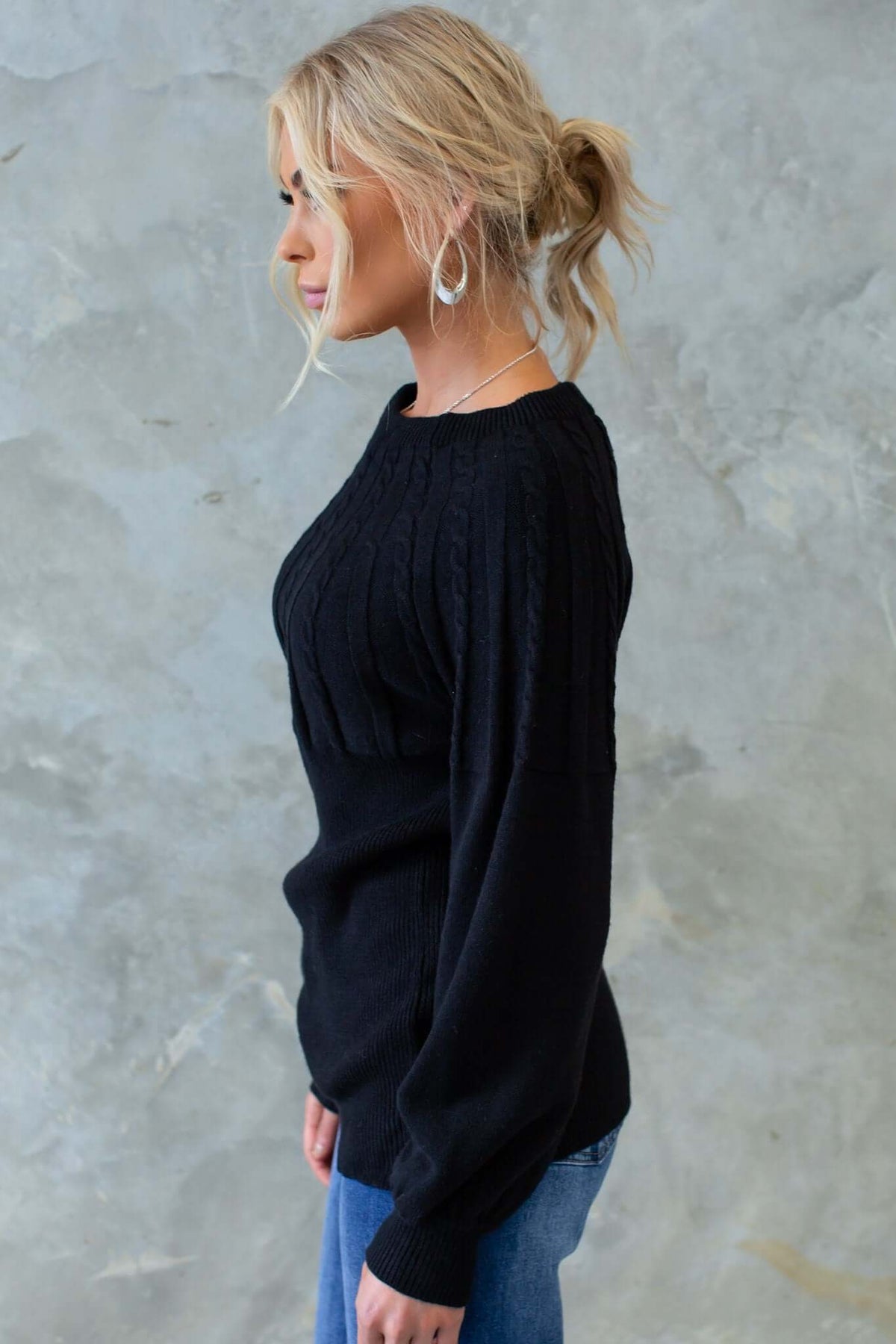 Profile view of a woman wearing a black Alma jumper with a round neckline and cable knit design.