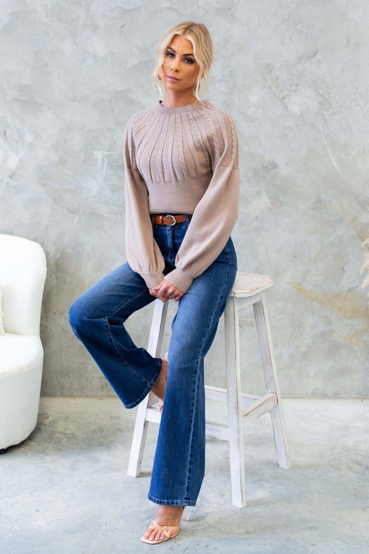 Alma Jumper with round neckline and cable knit style, paired with flared jeans and a belt, displayed in a relaxed setting.