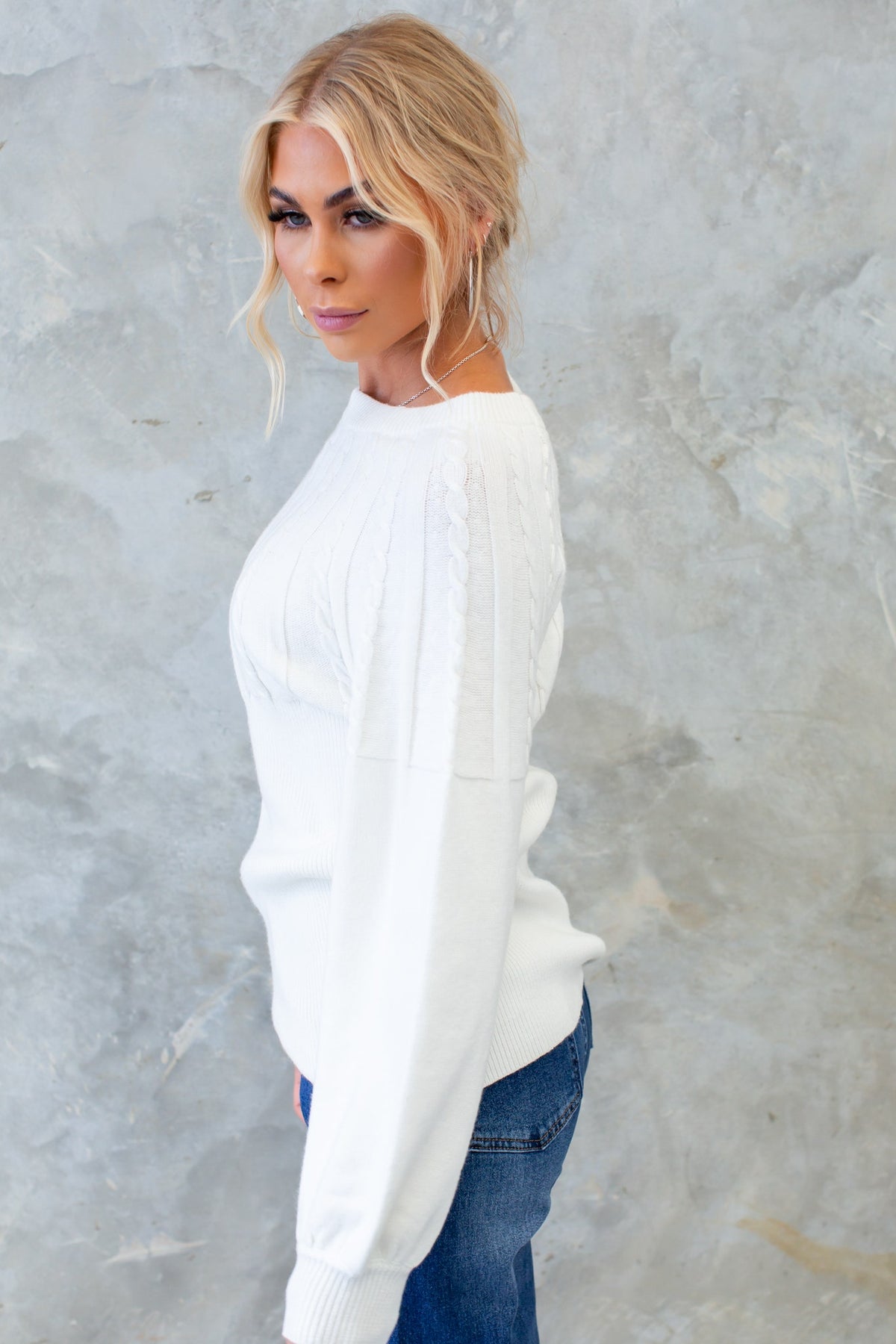 Side view of a woman wearing a white cable knit Alma Jumper, styled with relaxed-fit jeans.