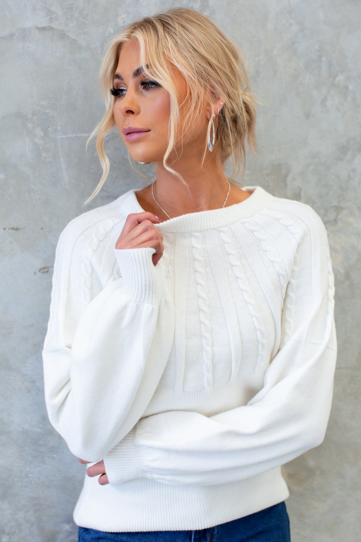Model wearing a white cable knit Alma Jumper with round neckline against a textured gray background.