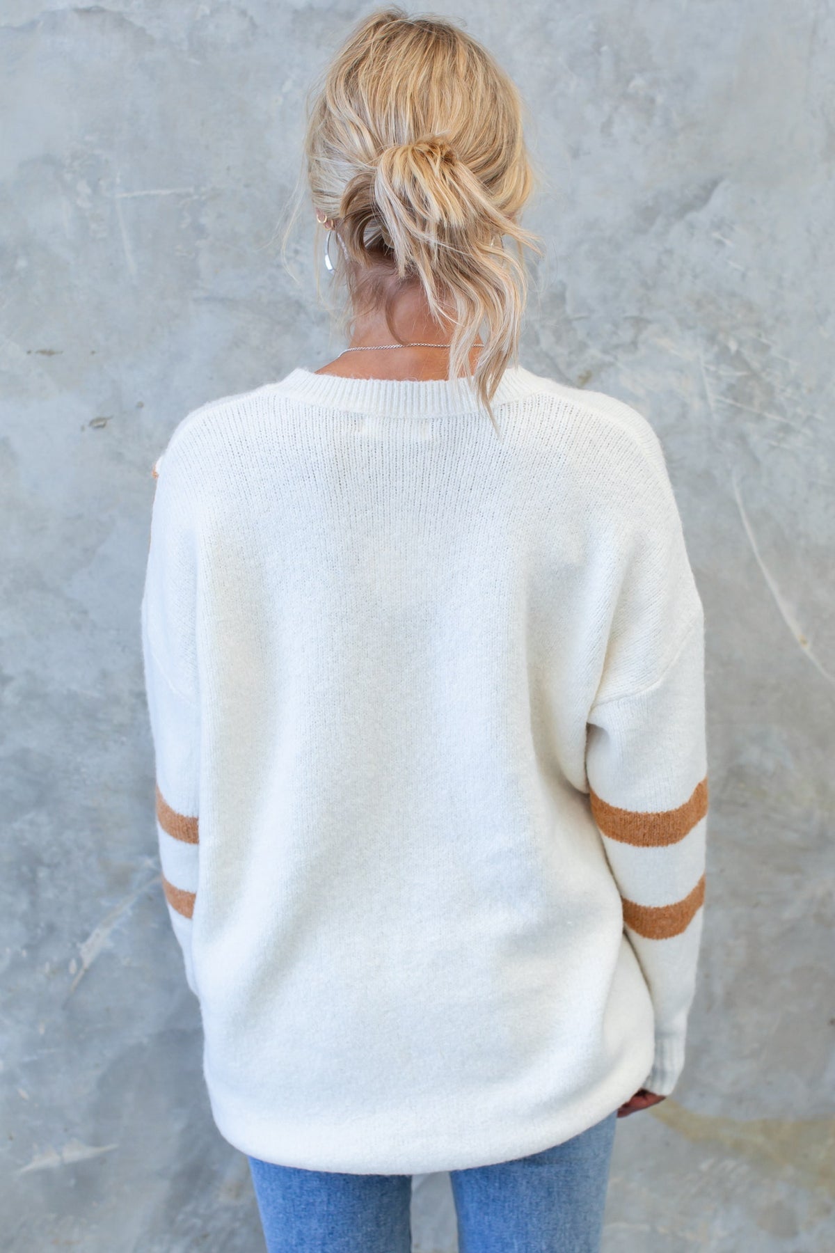 Back view of a woman wearing a cozy cream Wezen Jumper with brown stripes and relaxed fit, paired with blue jeans.